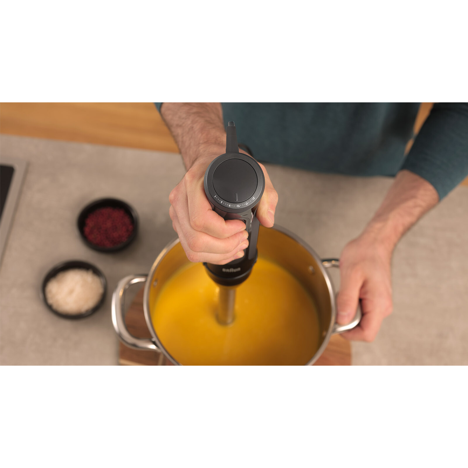 A person using a Braun hand blender in a pot. Yellow liquid is inside the pot.