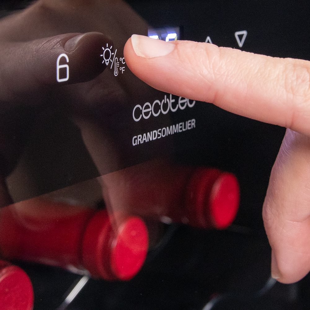 Un dedo toca el botón de luz en el panel de control de una vinoteca. Botellas de vino tinto visibles.
