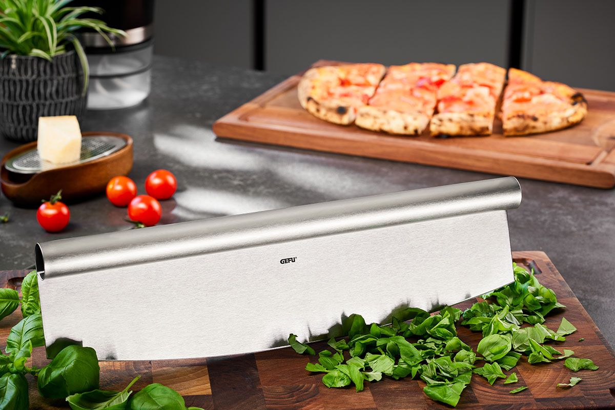 Pizzaschneider mit Basilikum auf einem Holzbrett. Pizza auf einem Holzbrett im Hintergrund, Tomaten, Käse und eine Pflanze sind ebenfalls vorhanden.