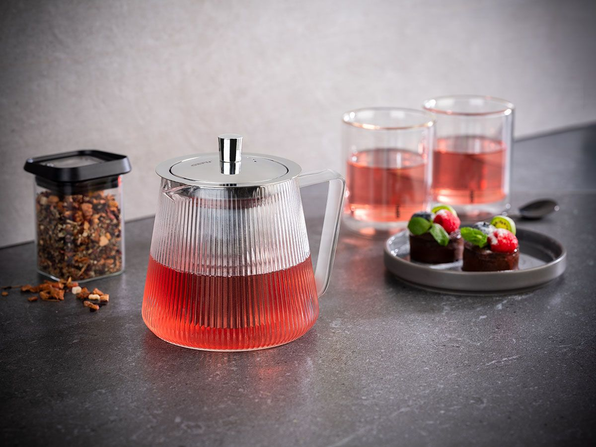Gläserne Teekanne mit roter Flüssigkeit, neben zwei Gläsern rotem Getränk und Kuchen.