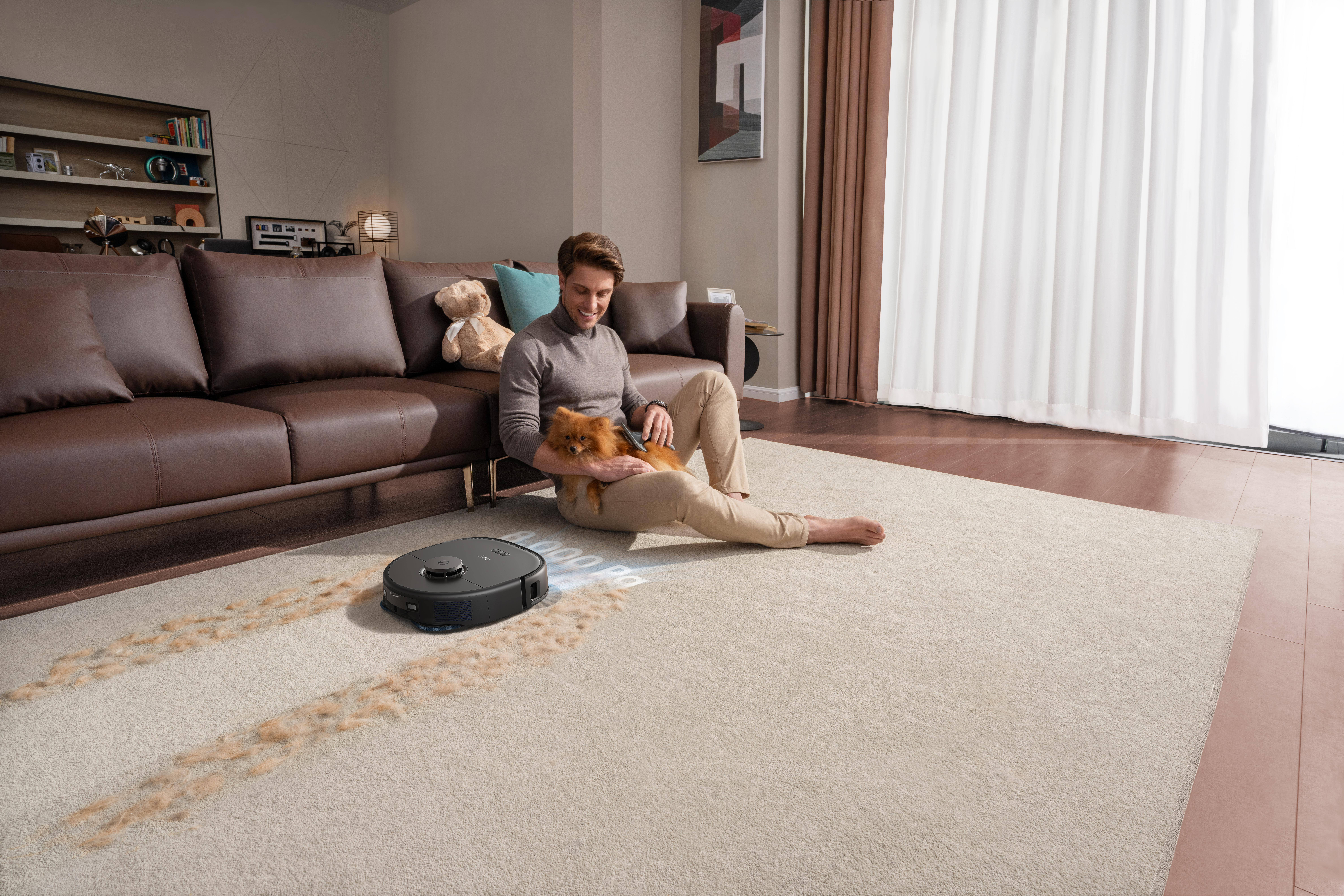 Mann sitzt auf dem Boden und streichelt einen Hund. Ein Roboter-Staubsauger reinigt einen Teppich mit Haaren, in der Nähe eines braunen Sofas.