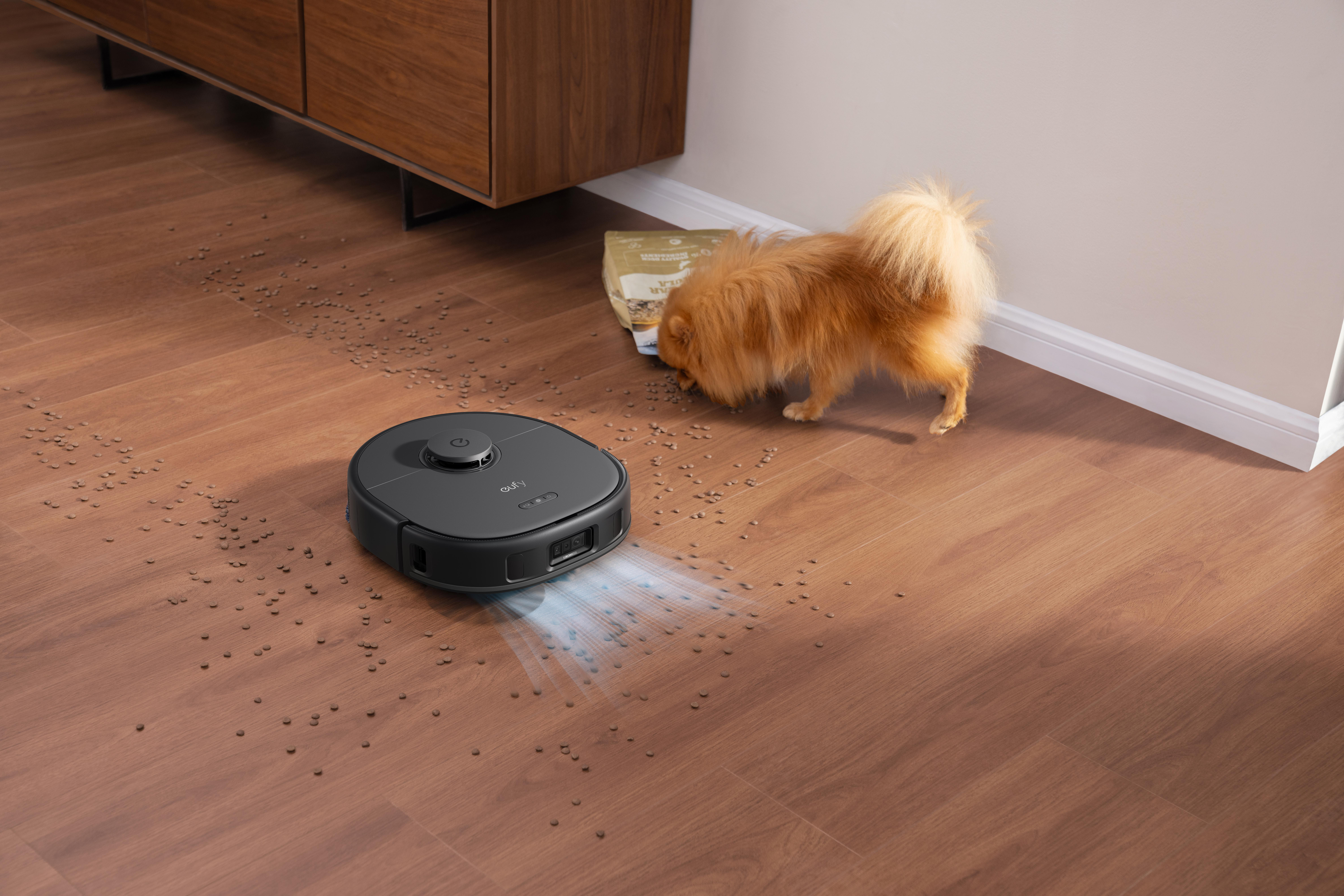 Ein Staubsaugerroboter reinigt verstreutes Tierfutter auf Holzboden, ein kleiner Hund neben der Futtersäcke.