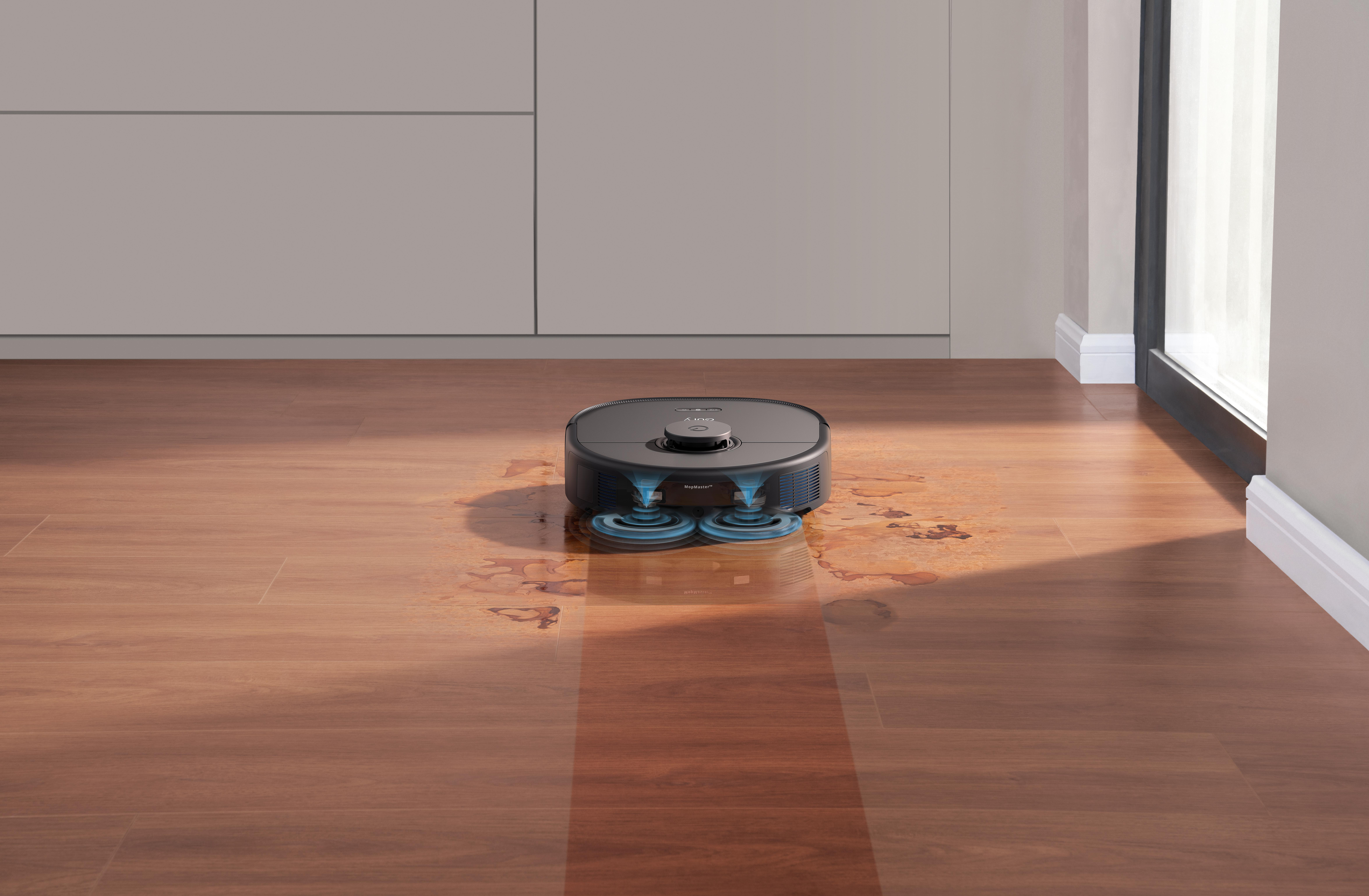 Ein Roboter wischt einen nassen Holzboden. Im Hintergrund sind ein Fenster und Schränke. Licht reflektiert sich auf dem Boden.