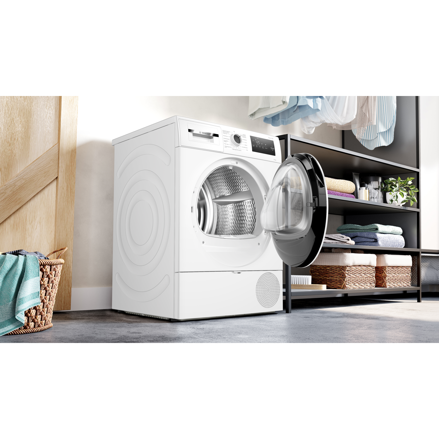 Een witte wasmachine in een wasruimte, deur open. Een mand met kleren links.