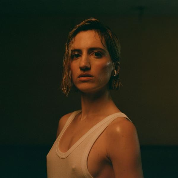 Frau in weißem Tanktop, kurzem blondem Haar, blickt in die Kamera. Dunkler Hintergrund.