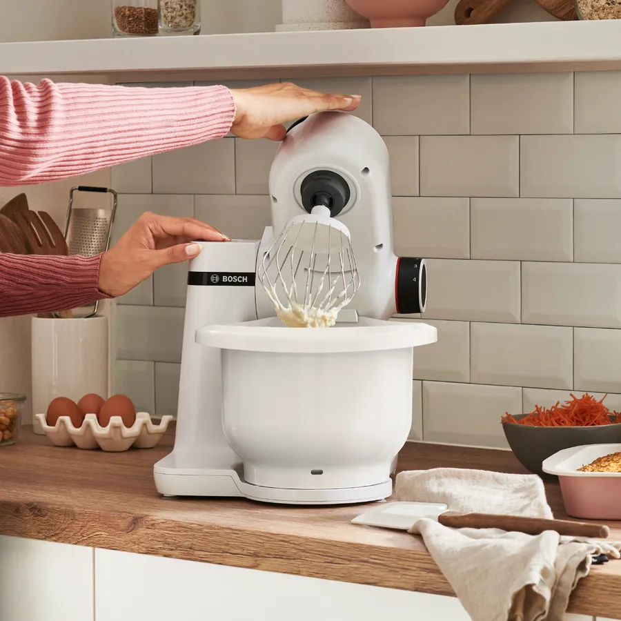 Robot pâtissier blanc, une personne ajoute des ingrédients, d'autres ingrédients sont visibles.