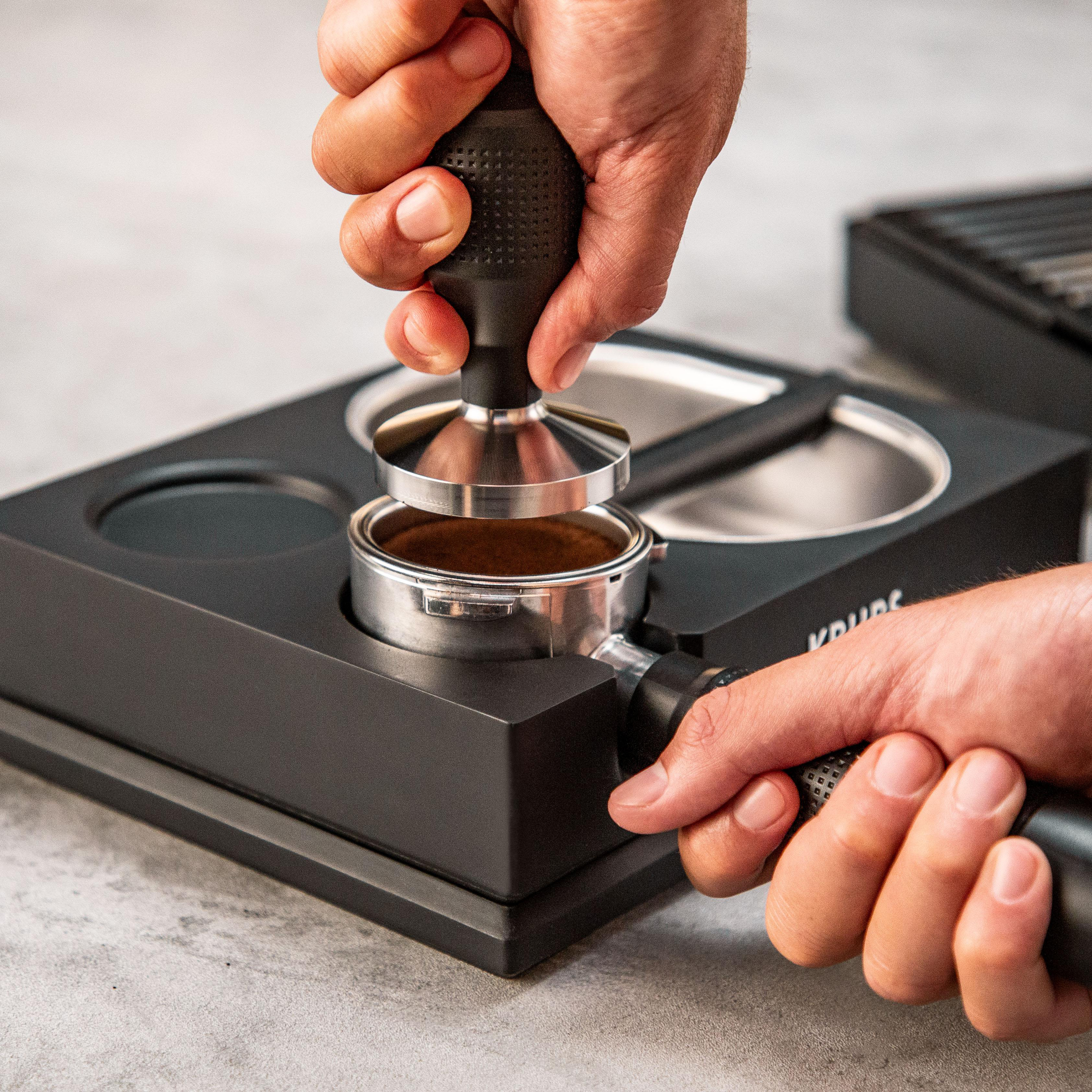 Hände drücken einen Tamper in einen mit Kaffeepulver gefüllten Siebträger.