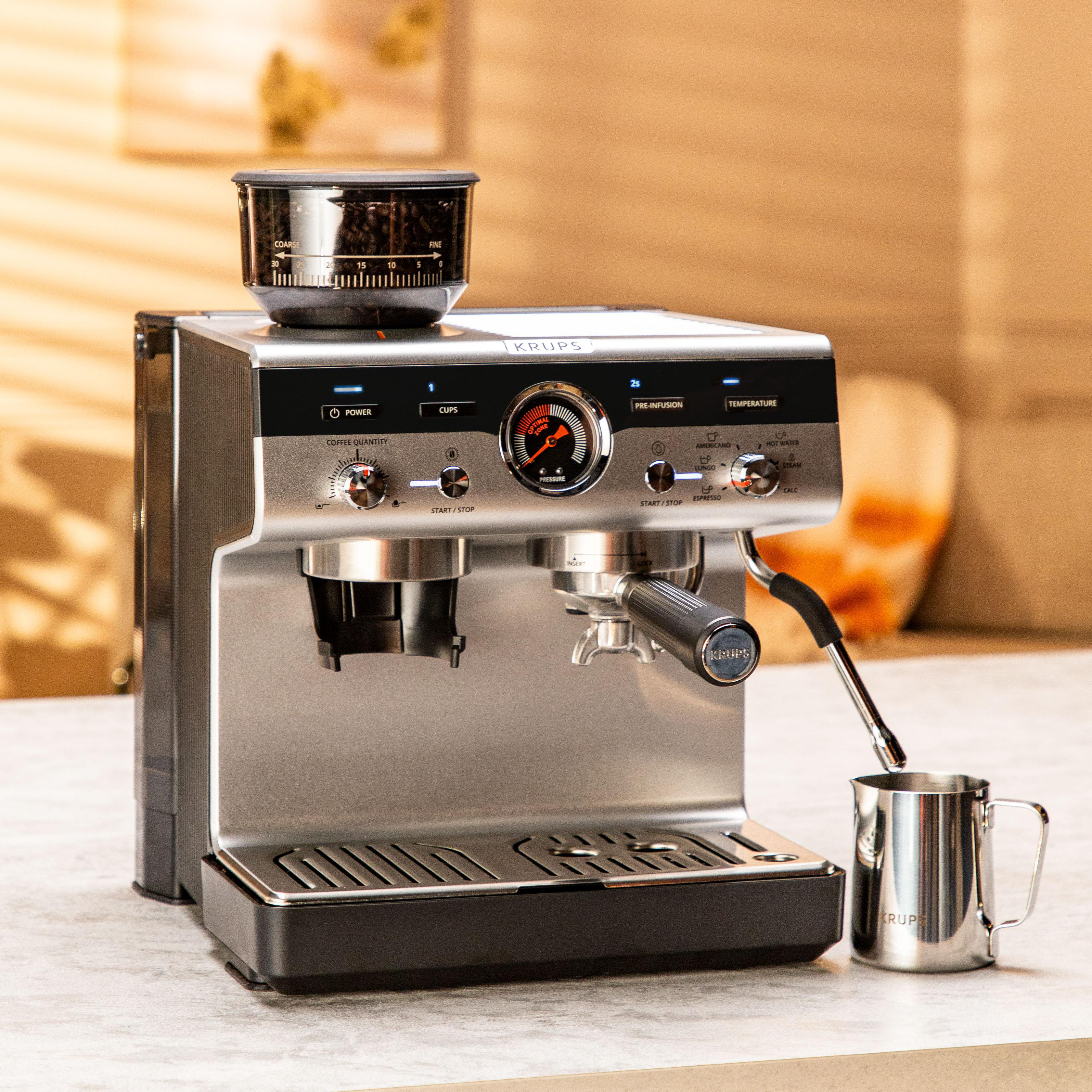 Silberne Espressomaschine mit Mahlwerk, Bedienelementen und Milchaufschäumdüse. Ein Metallbecher steht unter der Düse.