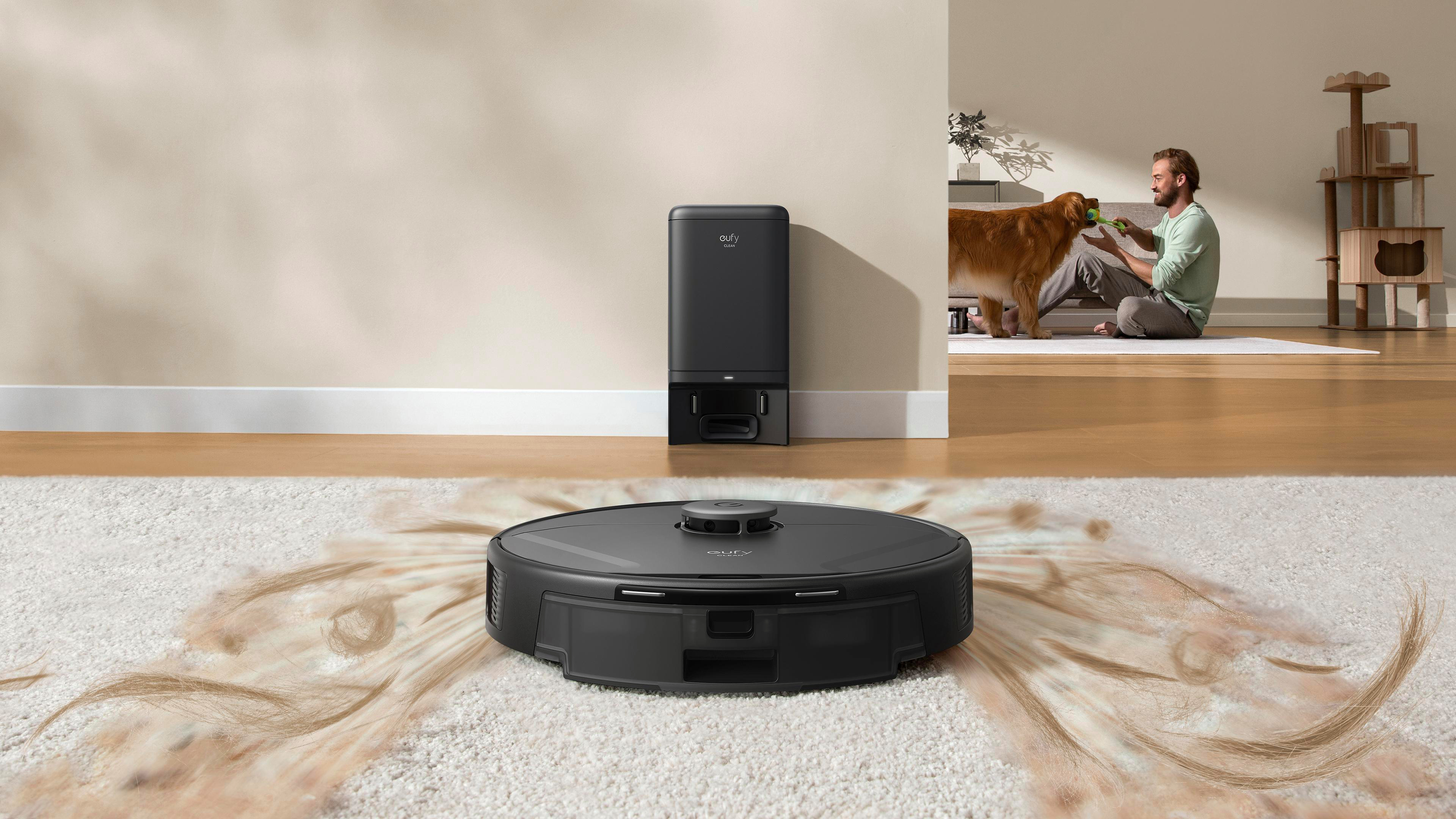 Roboter-Staubsauger auf einem Teppich, eine Ladestation, ein Mann, der mit einem Hund spielt, und ein Katzenbaum im Hintergrund.