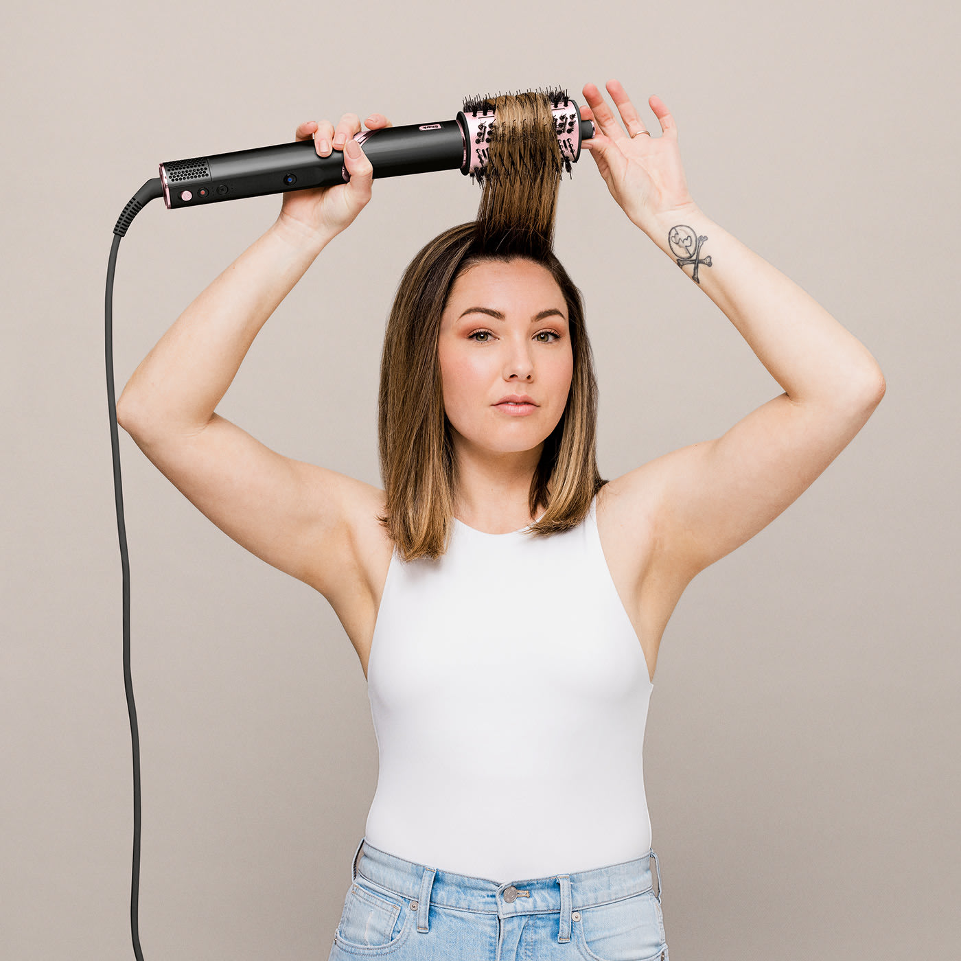 Eine Frau benutzt ein Haarstyling-Gerät. Sie trägt ein weißes Tanktop und Jeans. Ein Tattoo ist auf ihrem Arm sichtbar.