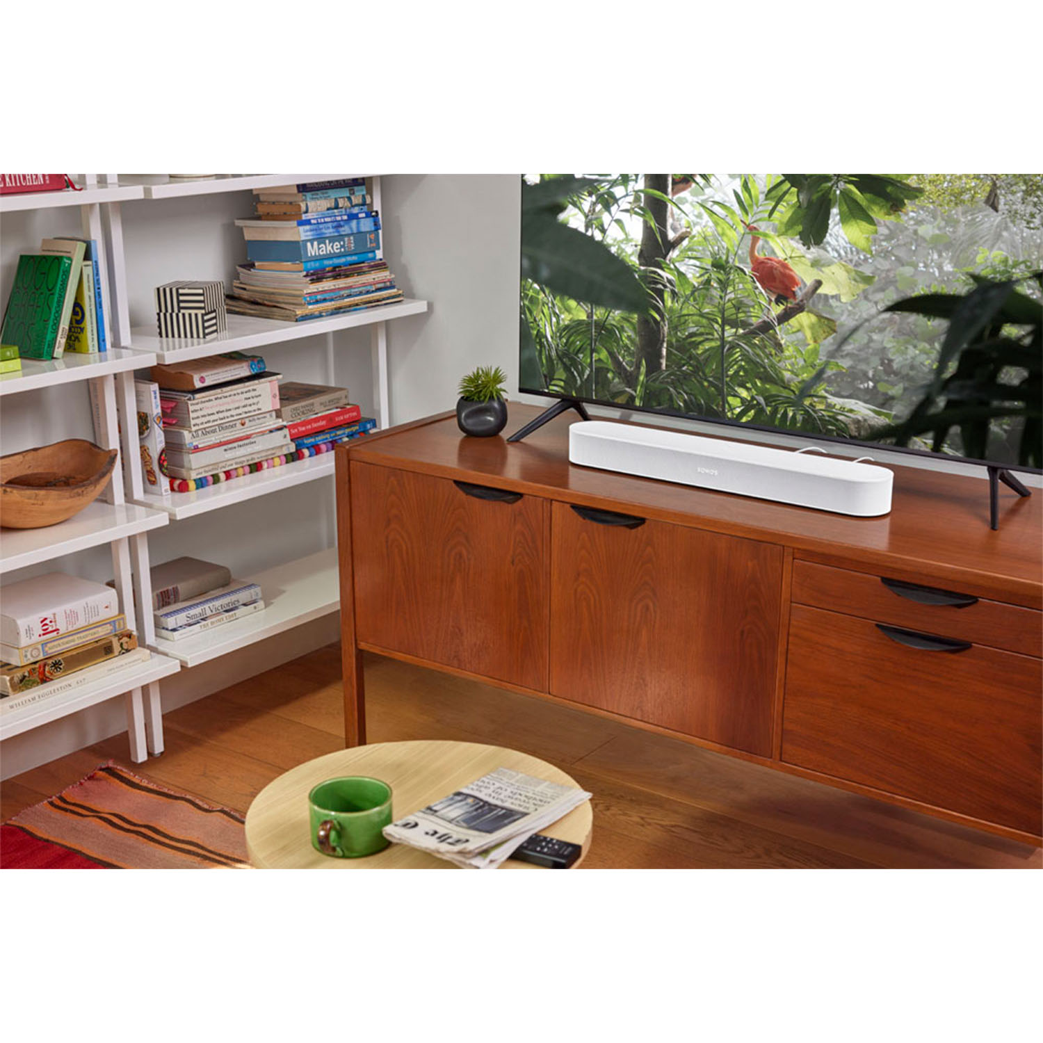 Una soundbar bianca è posizionata su un mobile in legno di fronte a un televisore. Librerie e una pianta sullo sfondo.