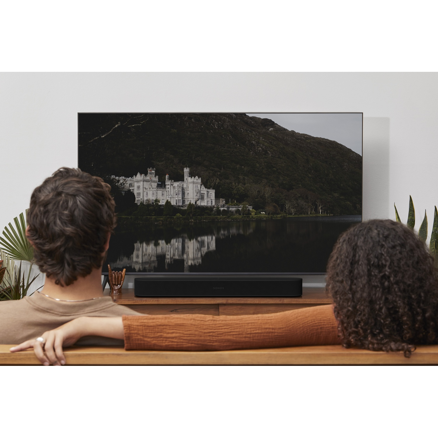 Due persone guardano la TV. Una soundbar nera è posizionata sotto la TV. Lo sfondo è un lago e un castello.