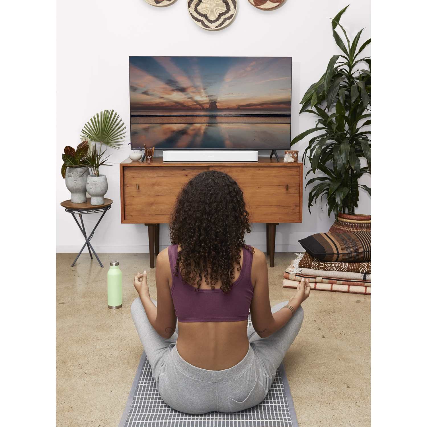 Donna fa yoga sul tappetino, guarda la TV, con piante e borraccia.