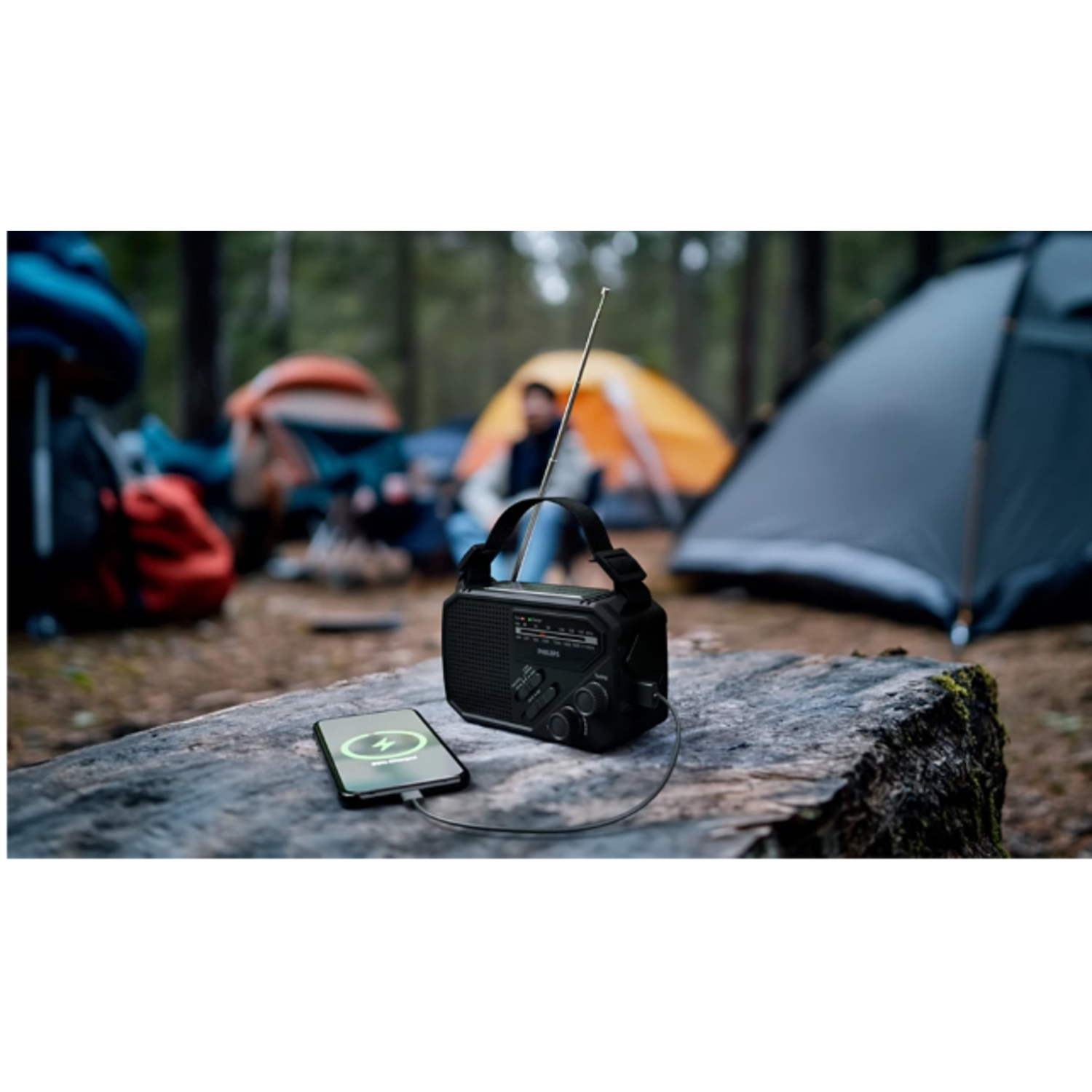 Een zwarte radio die een telefoon oplaadt op een houten stam in een bos, met tenten op de achtergrond.