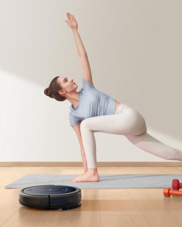 Frau macht Yoga neben einem Roboterstaubsauger. Graue Matte, Holzboden, rote Gewichte. Hellblaues Oberteil, weiße Hose.