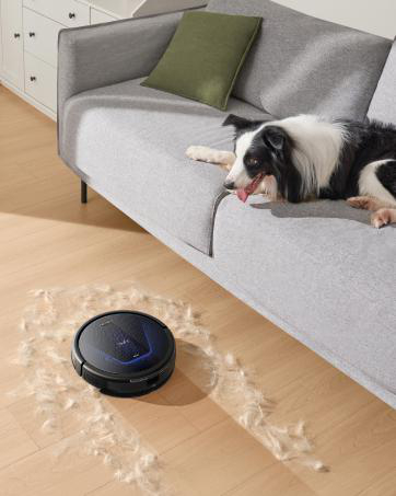 Ein schwarzer Roboterstaubsauger reinigt Hundehaare in der Nähe einer Couch, auf der ein schwarz-weißer Hund ruht.
