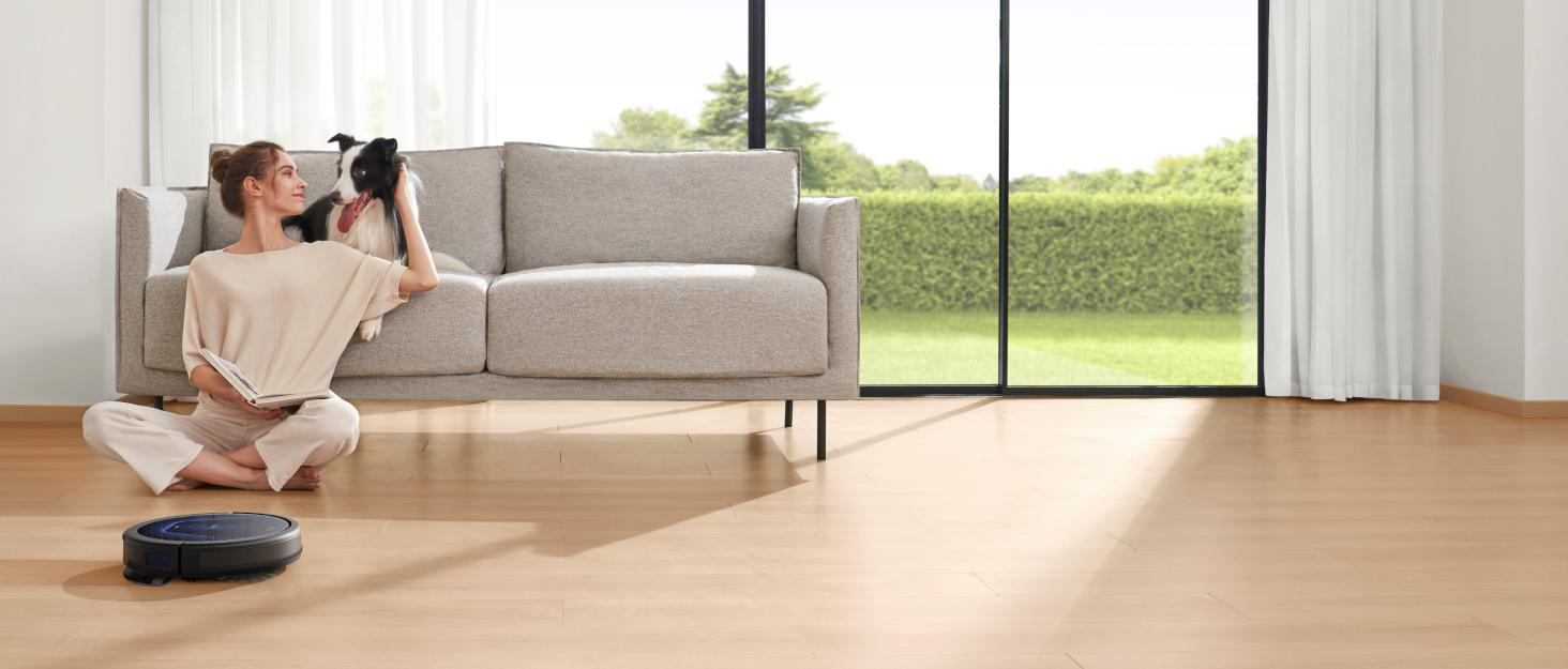 Eine Frau sitzt mit einem Hund auf einem Sofa. Ein Roboterstaubsauger reinigt einen Holzboden. Draußen sind Rasen und Bäume zu sehen.