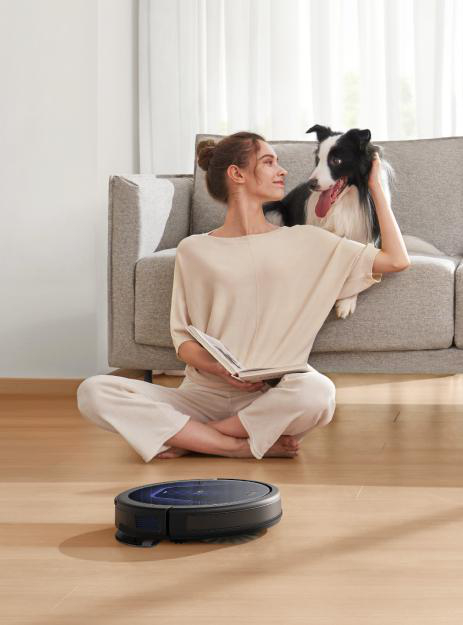 Frau liest mit Hund ein Buch. Ein Roboter-Staubsauger ist im Vordergrund. Der Hintergrund ist ein Wohnzimmer.