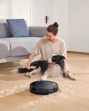 Eine Frau bürstet einen schwarz-weißen Hund auf dem Boden. Ein Roboterstaubsauger befindet sich neben dem Hund. Im Hintergrund ist ein graues Sofa zu sehen.