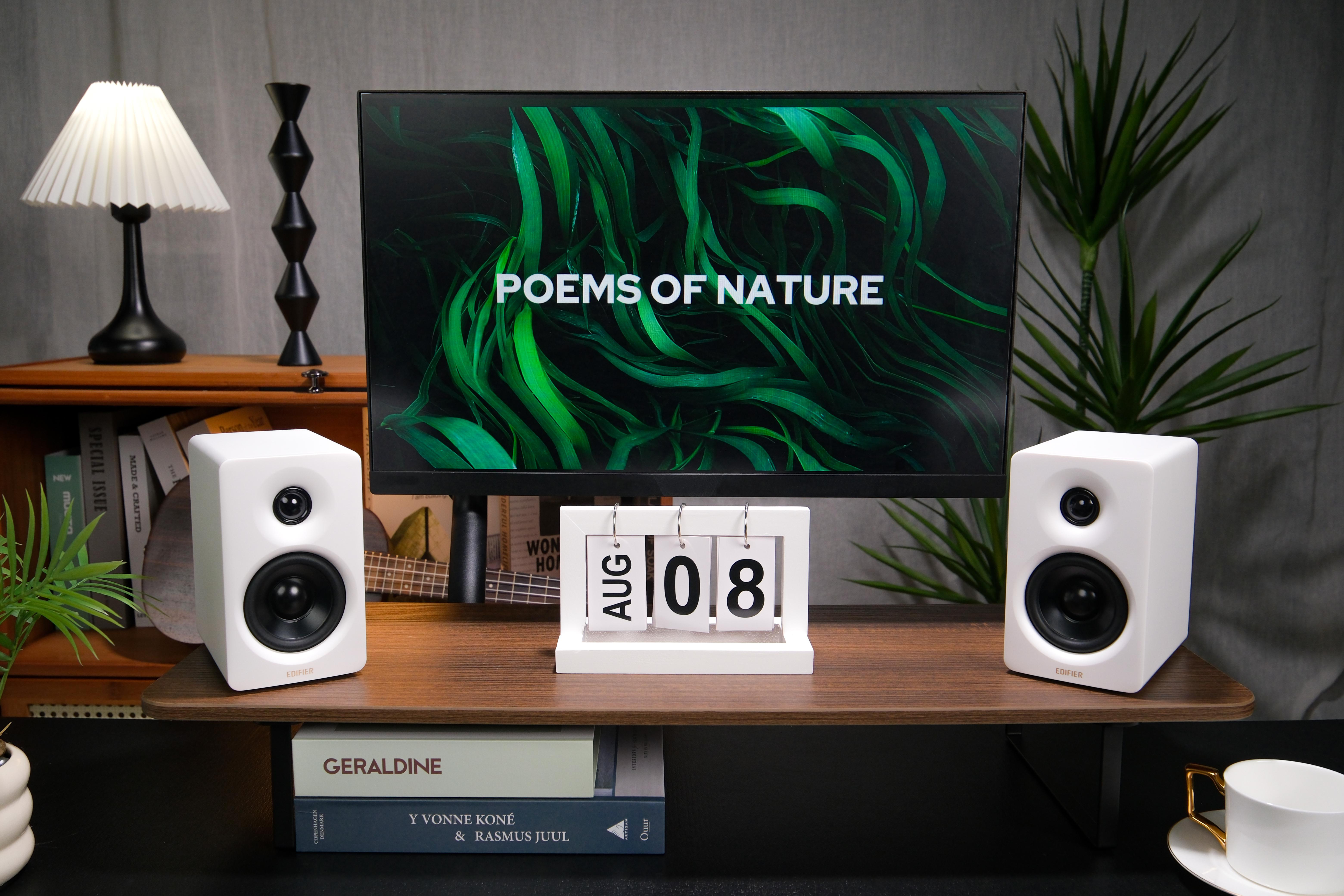 Schreibtisch mit Monitor, Lautsprechern und Kalender mit 08. August. Bücher und Lampe vorhanden.