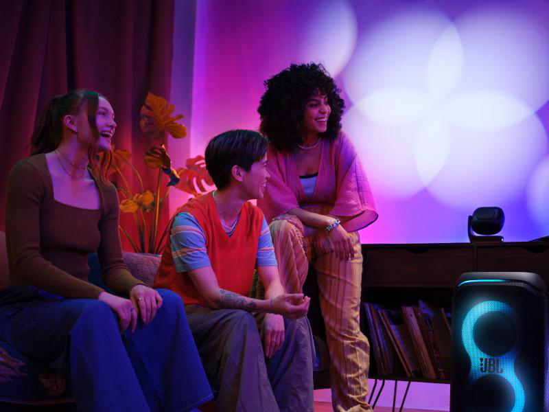 Drei lachende Personen in einem Raum mit einem JBL-Lautsprecher und farbenfroher Ambientebeleuchtung.