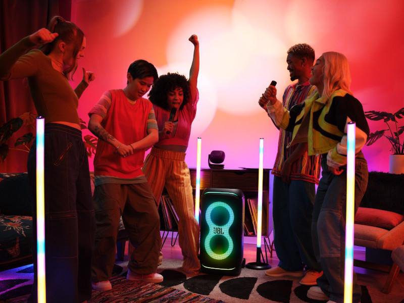 Menschen tanzen und singen um einen JBL-Lautsprecher mit Lichtsäulen in einem Raum mit farbenfroher Beleuchtung.