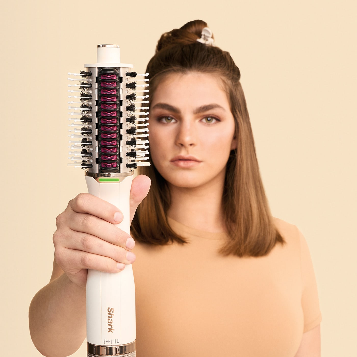 Una donna tiene in mano un arricciacapelli bianco e nero. I suoi capelli sono raccolti in uno chignon. Sfondo beige.