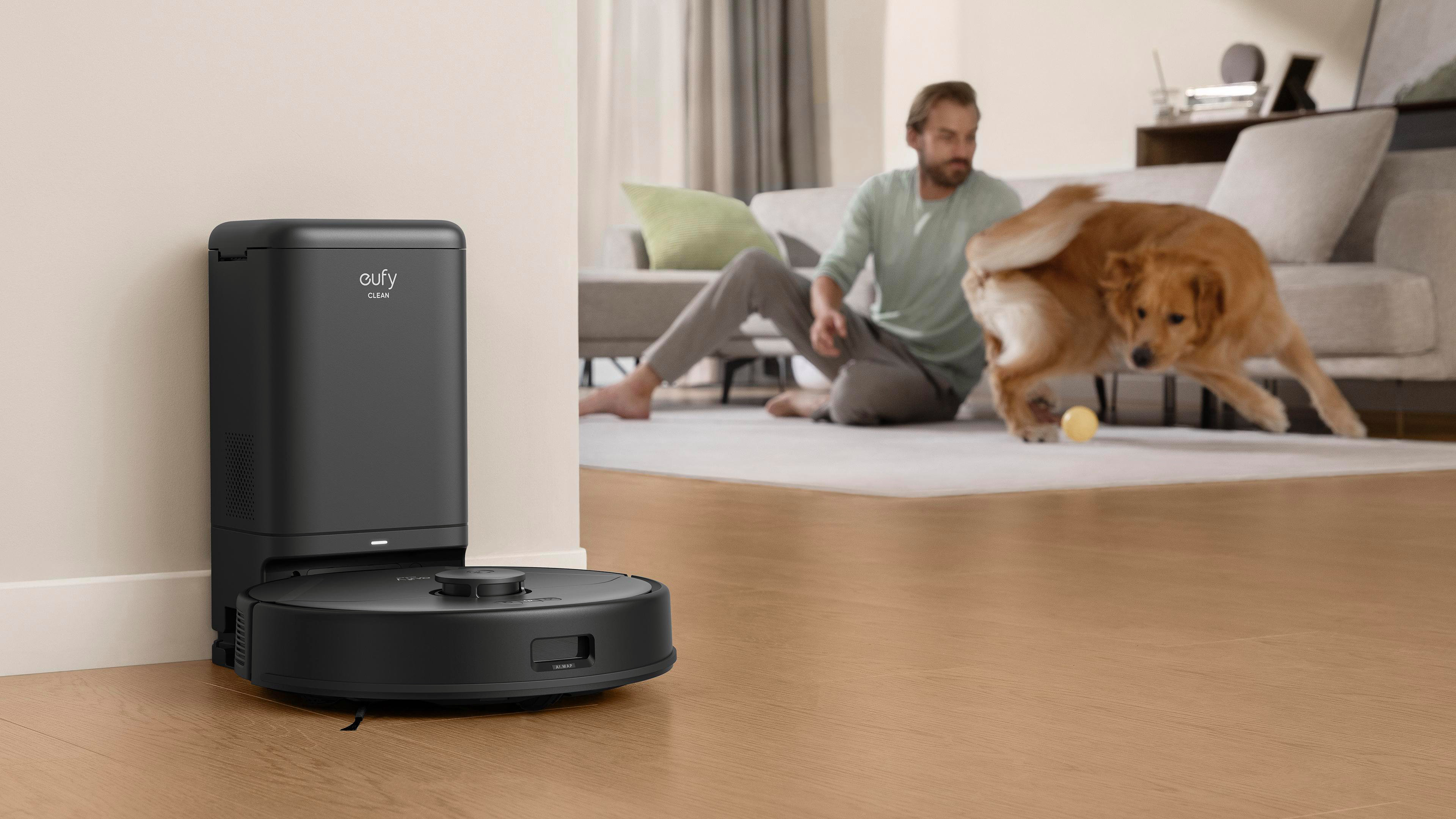 Ein schwarzer Roboter-Staubsauger steht an einer Wand. Im Hintergrund spielen ein Mann und ein Hund. Ein gelber Ball liegt auf dem Boden.