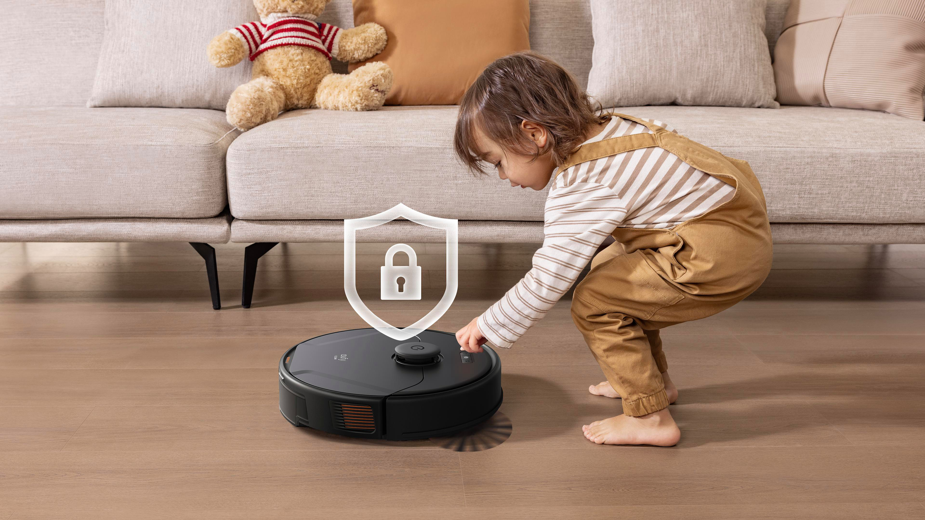 Ein Kind in Latzhosen interagiert mit einem schwarzen Roboter-Staubsauger. Ein Teddybär sitzt im Hintergrund auf einem Sofa.