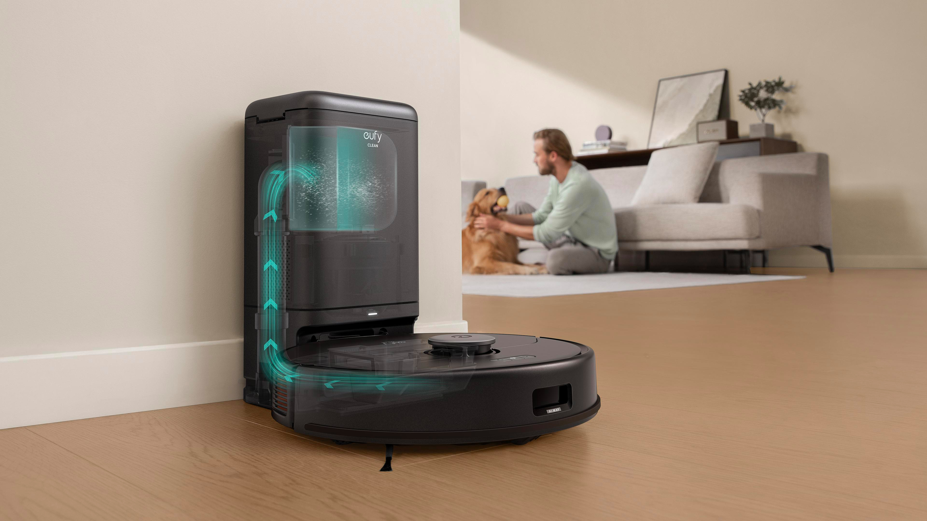 Ein Roboter-Staubsauger und seine Ladestation befinden sich im Vordergrund. Im Hintergrund spielt ein Mann mit einem Hund.