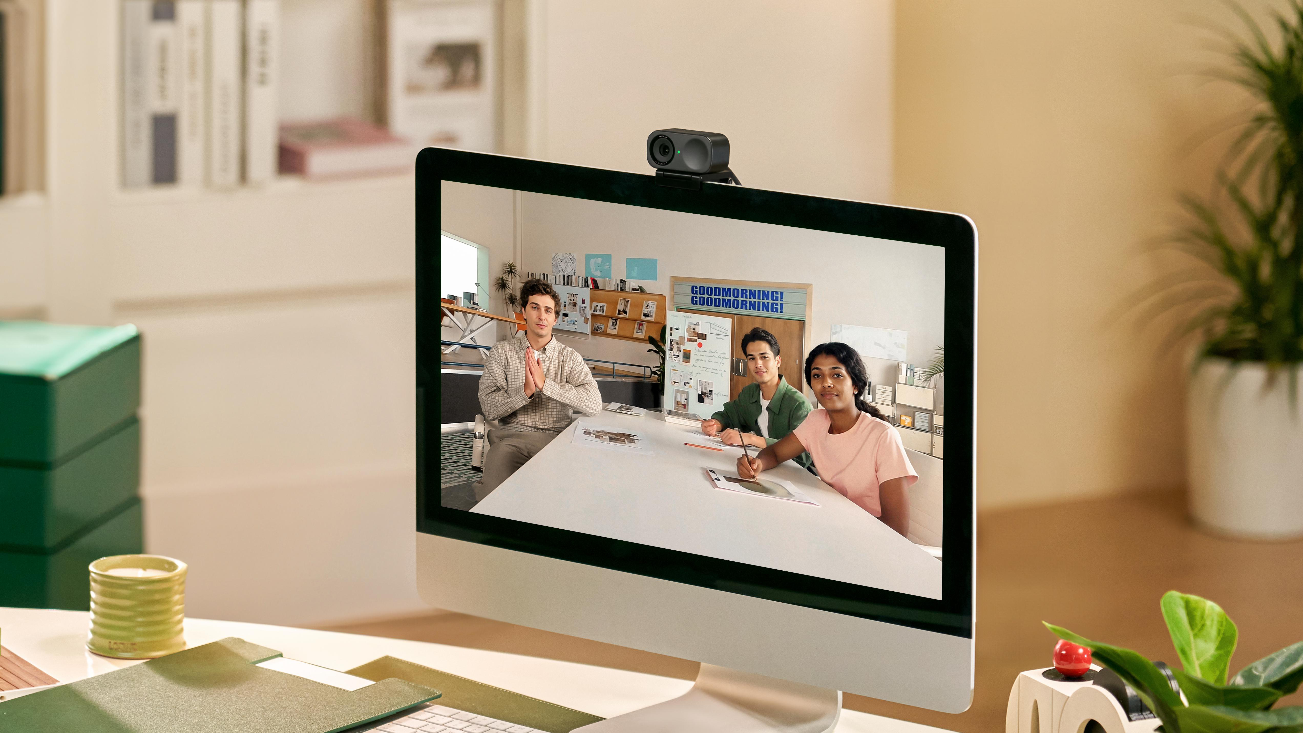 Bildschirm mit Personen, Webcam oben, Schreibtisch mit Gegenständen, Klassenzimmer.