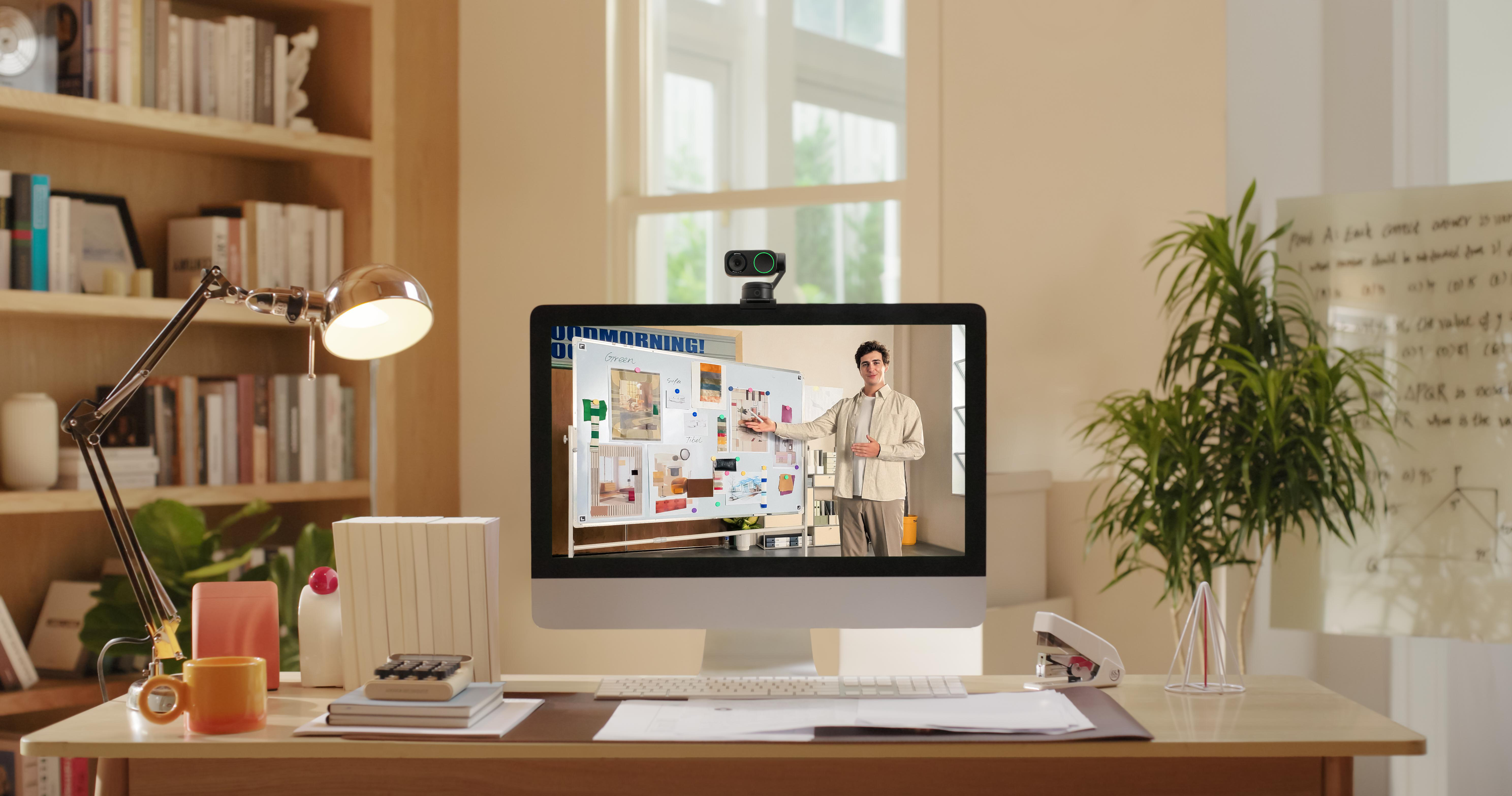 Person auf Bildschirm, Tafel mit Notizen, Webcam, Schreibtisch mit Gegenständen.