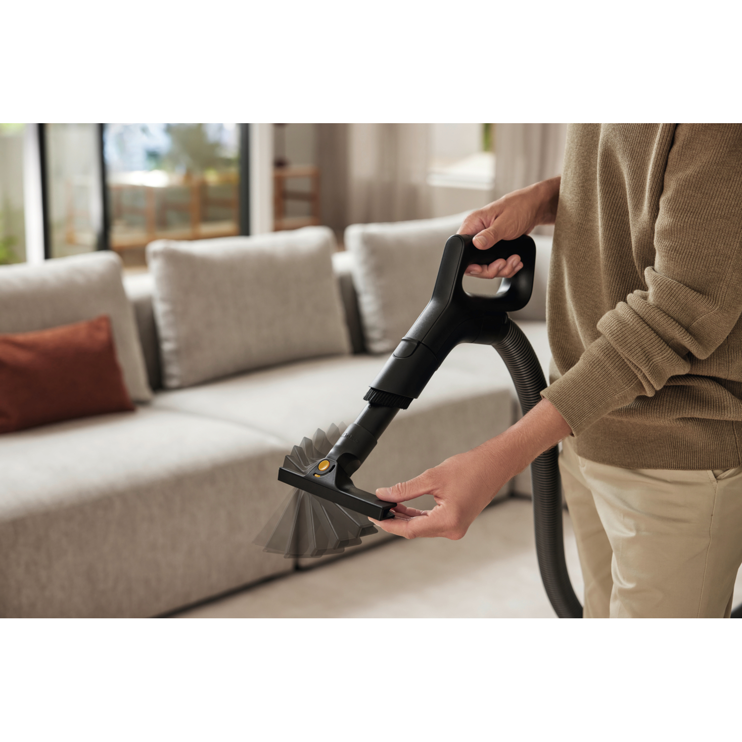 A person holds a vacuum cleaner attachment, cleaning a light-colored carpet in a living room.