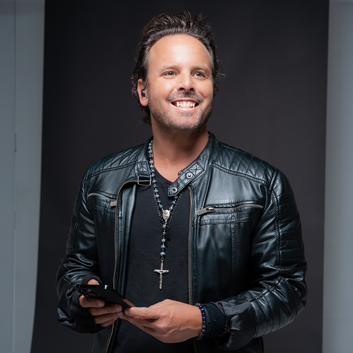 Smiling man wearing earbuds, holding phone, black leather jacket.