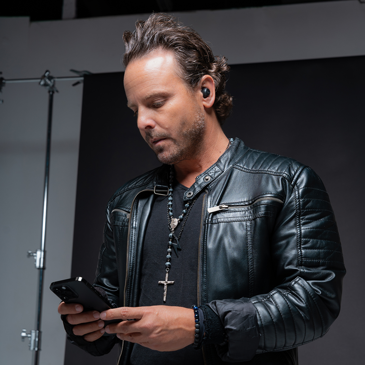 Man with earbuds looking at a phone. He wears a leather jacket and necklace. Studio background.
