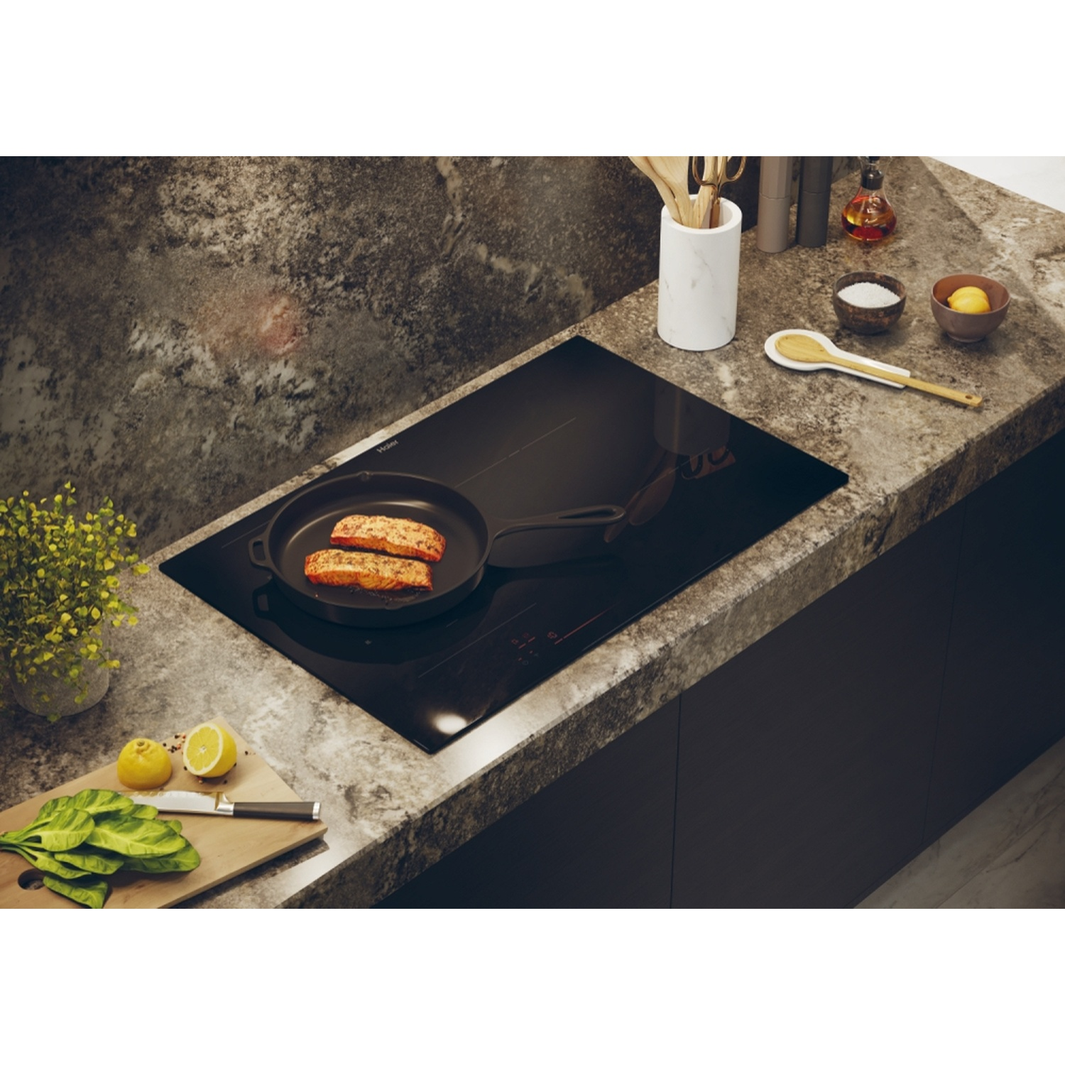 Piano cottura Haier nero con cibo in padella, limoni, erbe e utensili sul piano di lavoro.