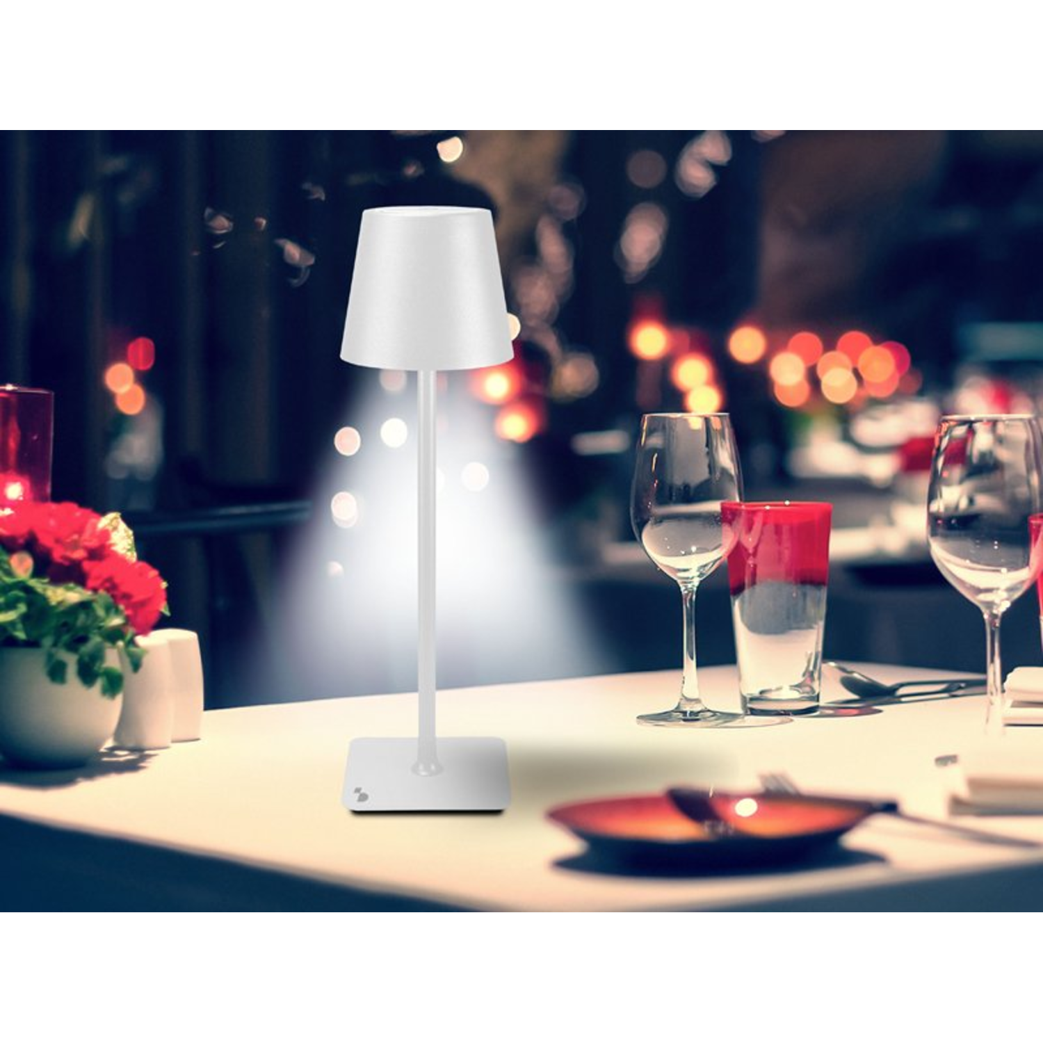 Witte tafellamp op een tafel met glazen en rode bloemen, verlicht door lamplicht.