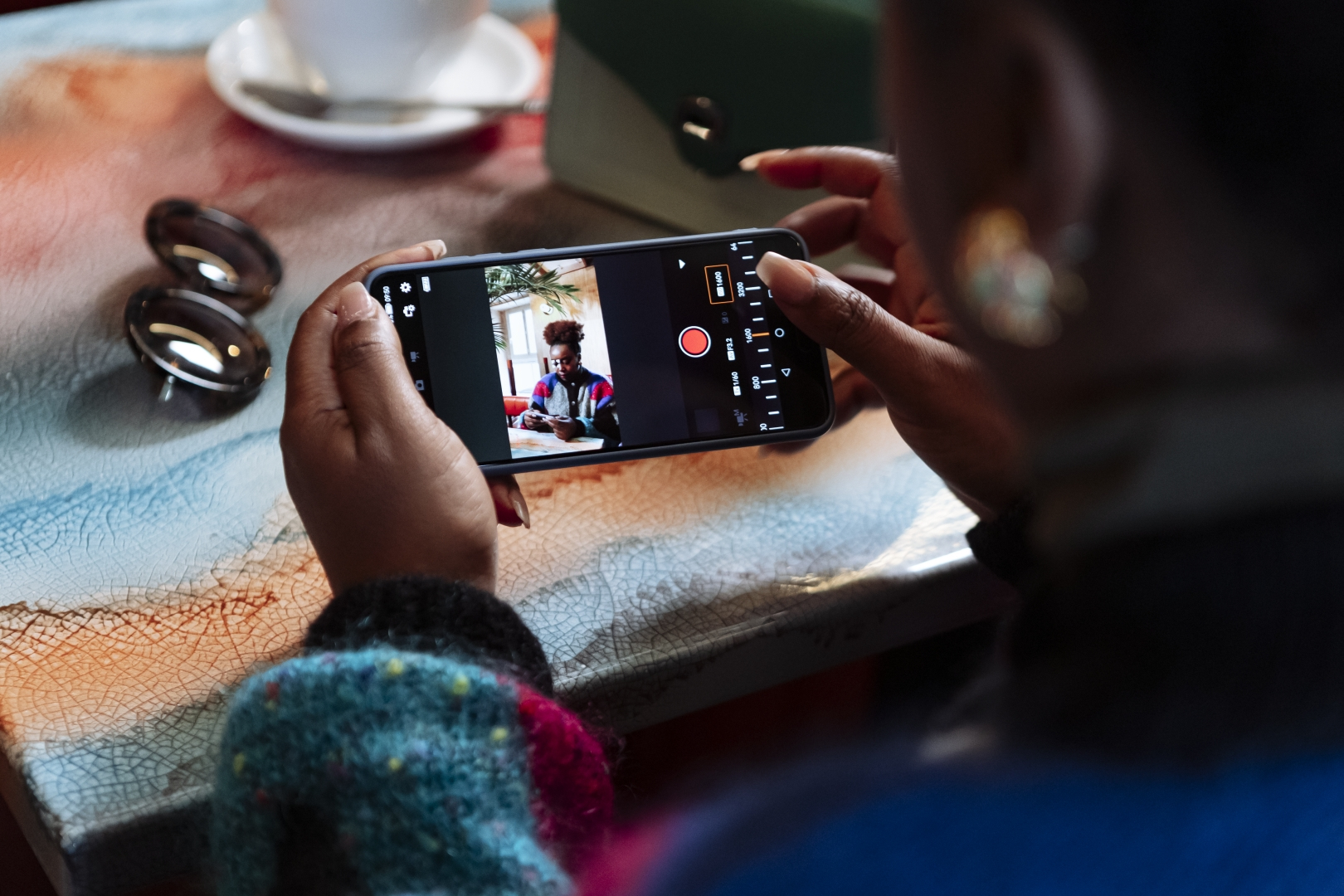 Eine Person hält ein Telefon mit einem Foto. Eine Kaffeetasse und Sonnenbrille liegen auf dem Tisch.