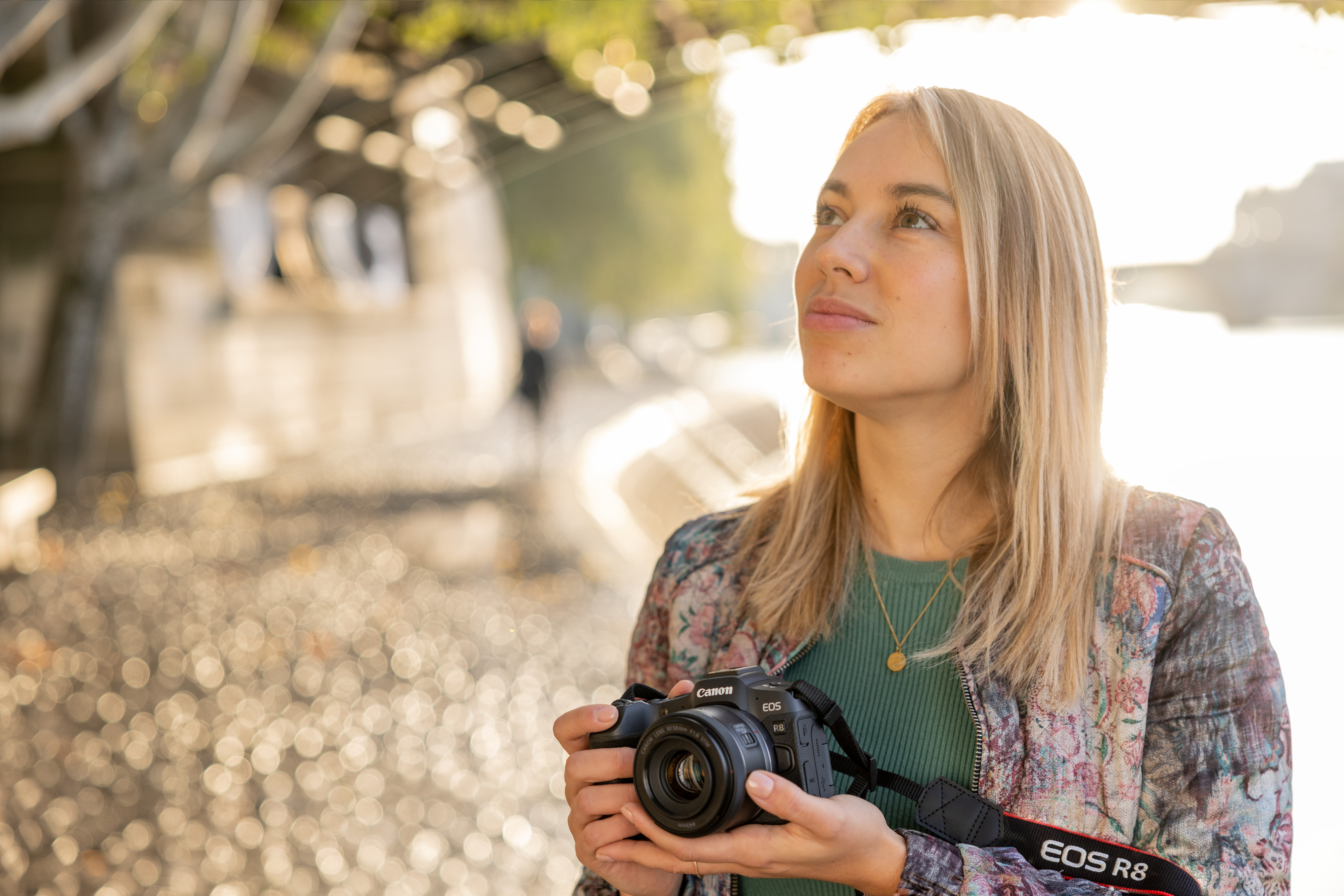 Blonde Frau hält Kamera, blickt nach oben. Sonniger Außenbereich mit Bokeh.