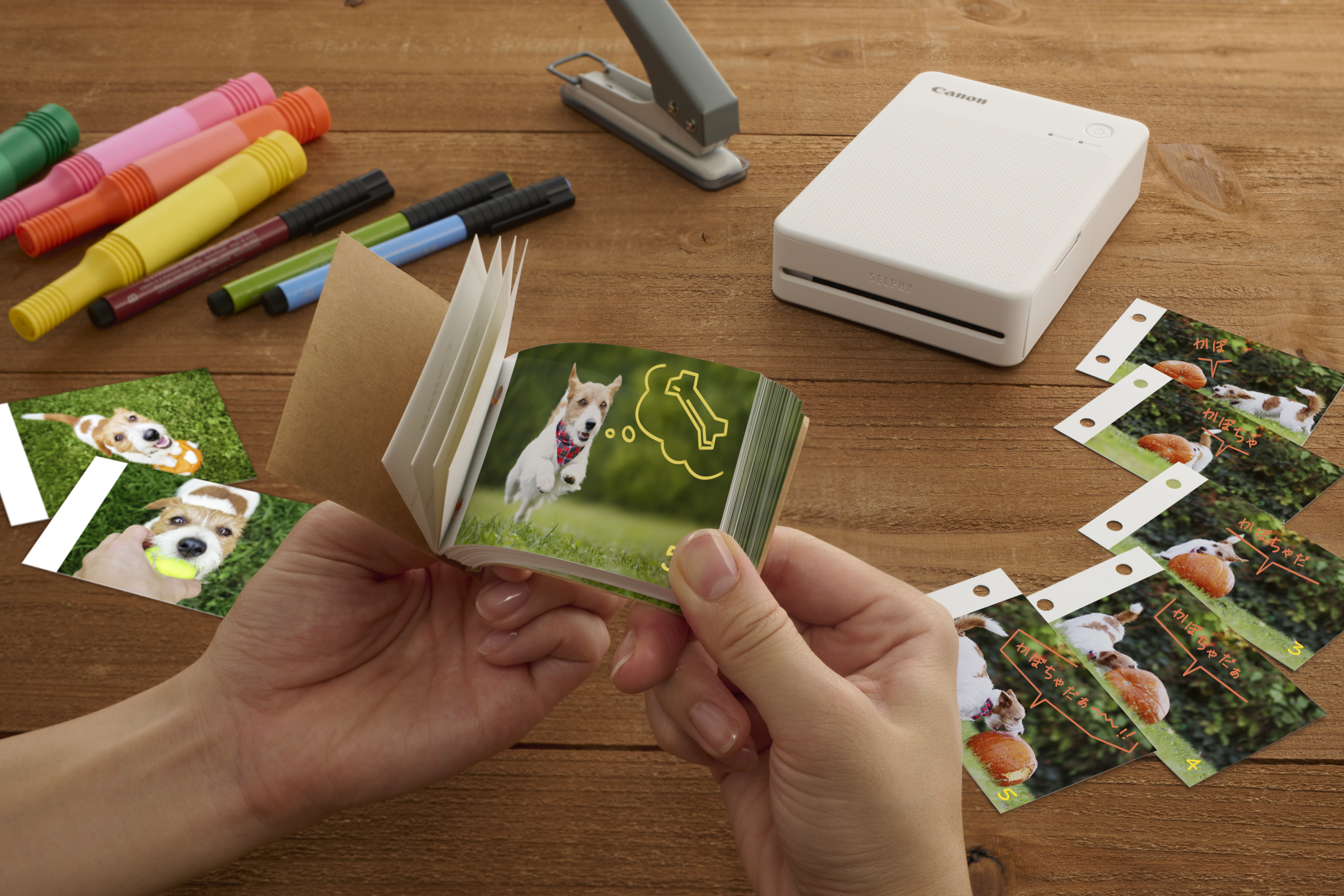 Person hält ein Mini-Fotoalbum und Fotos. Canon-Drucker. Marker und andere Gegenstände auf einem Holztisch.