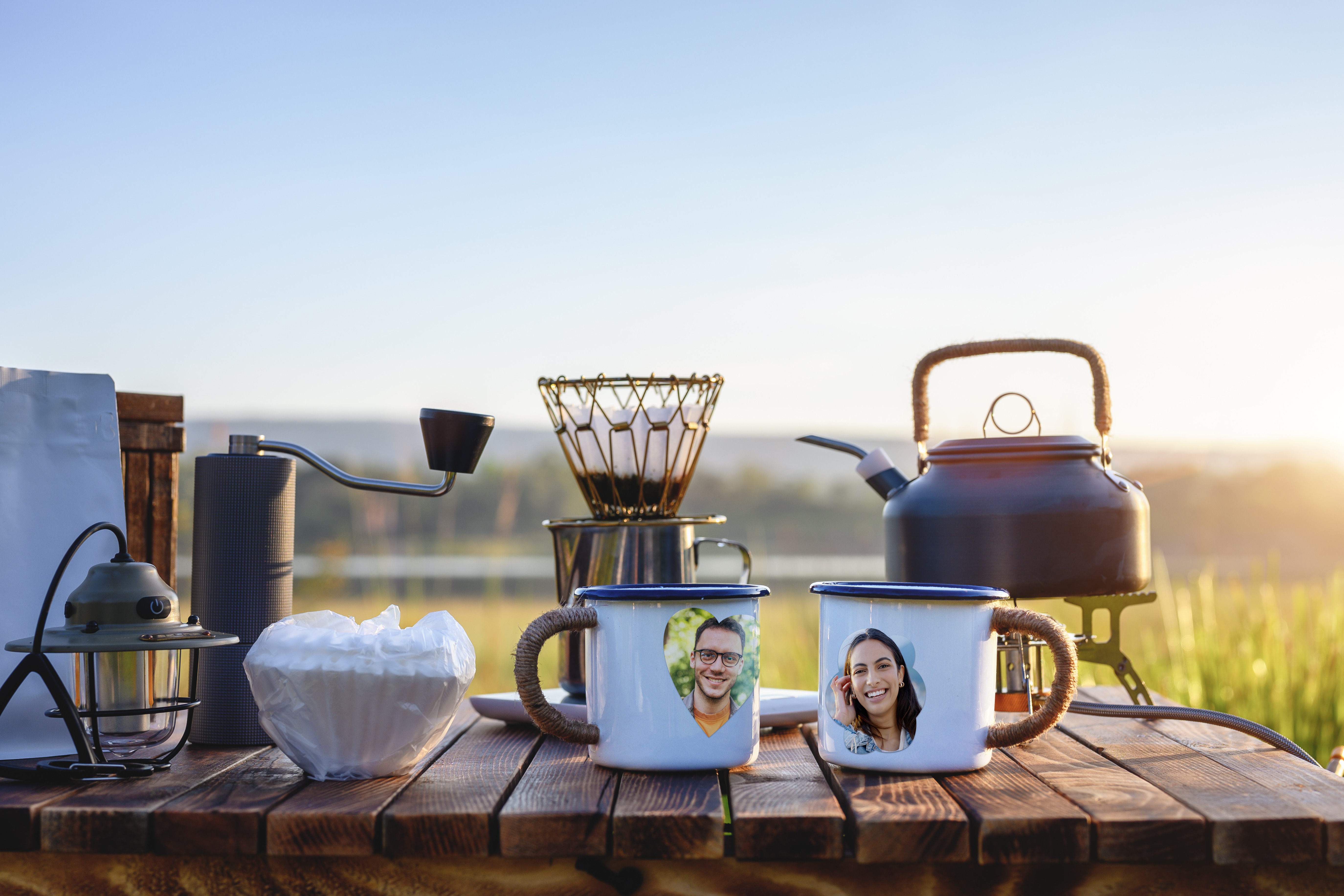 Zwei Tassen mit Fotos, Wasserkocher und andere Gegenstände auf einem Holztisch. Szene im Freien mit Sonnenlicht.