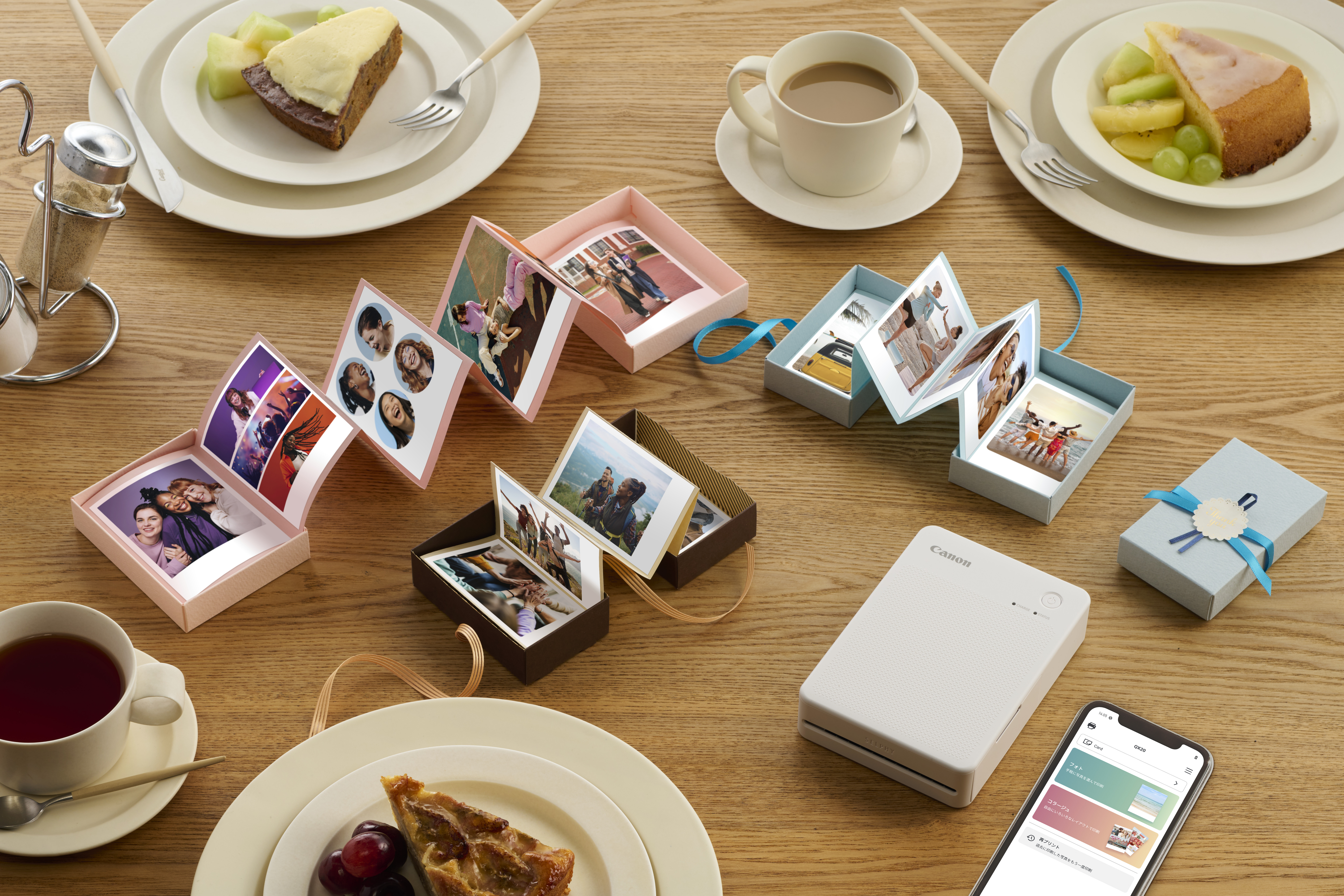 Fotos in kleinen, offenen Fotoboxen auf einem Holztisch. Kuchen, Getränke und ein Telefon mit App sichtbar.