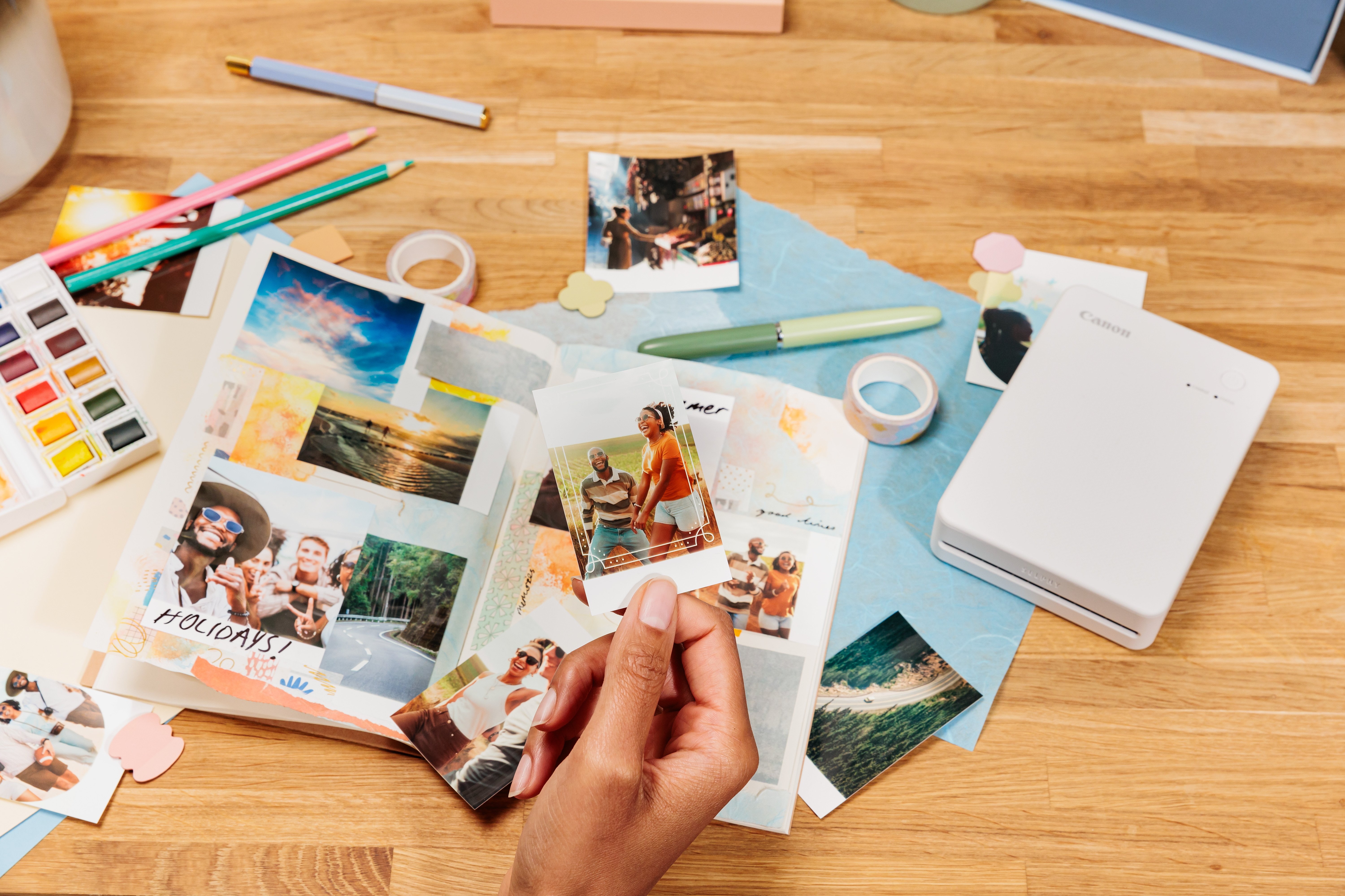 Eine Hand hält ein Foto in einem Album. Auf einem Holztisch liegen weitere Fotos, Bleistifte und ein Drucker.