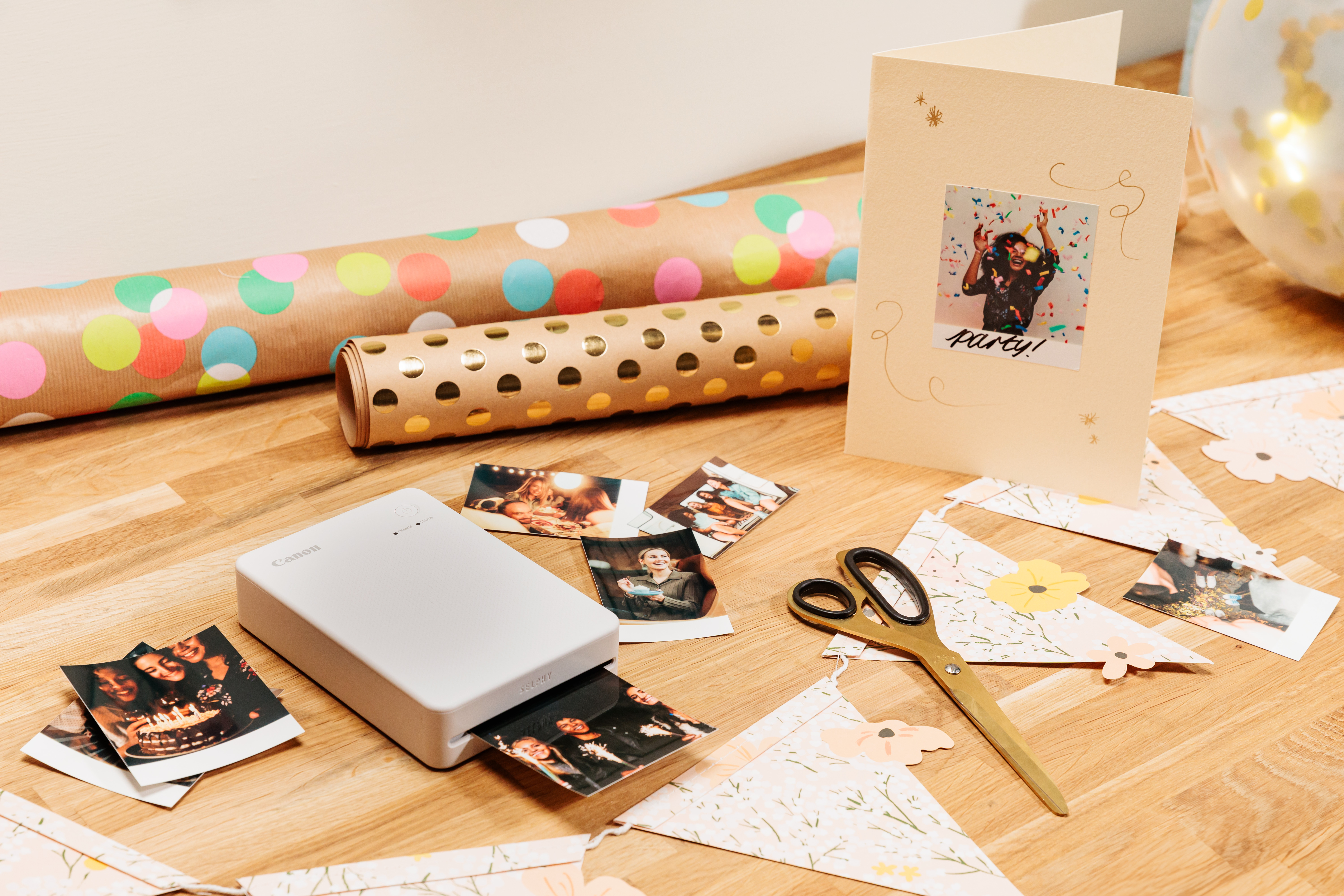 Ein tragbarer Drucker, Fotos und eine Geburtstagskarte liegen auf einem Holztisch mit Geschenkpapier und Schere.
