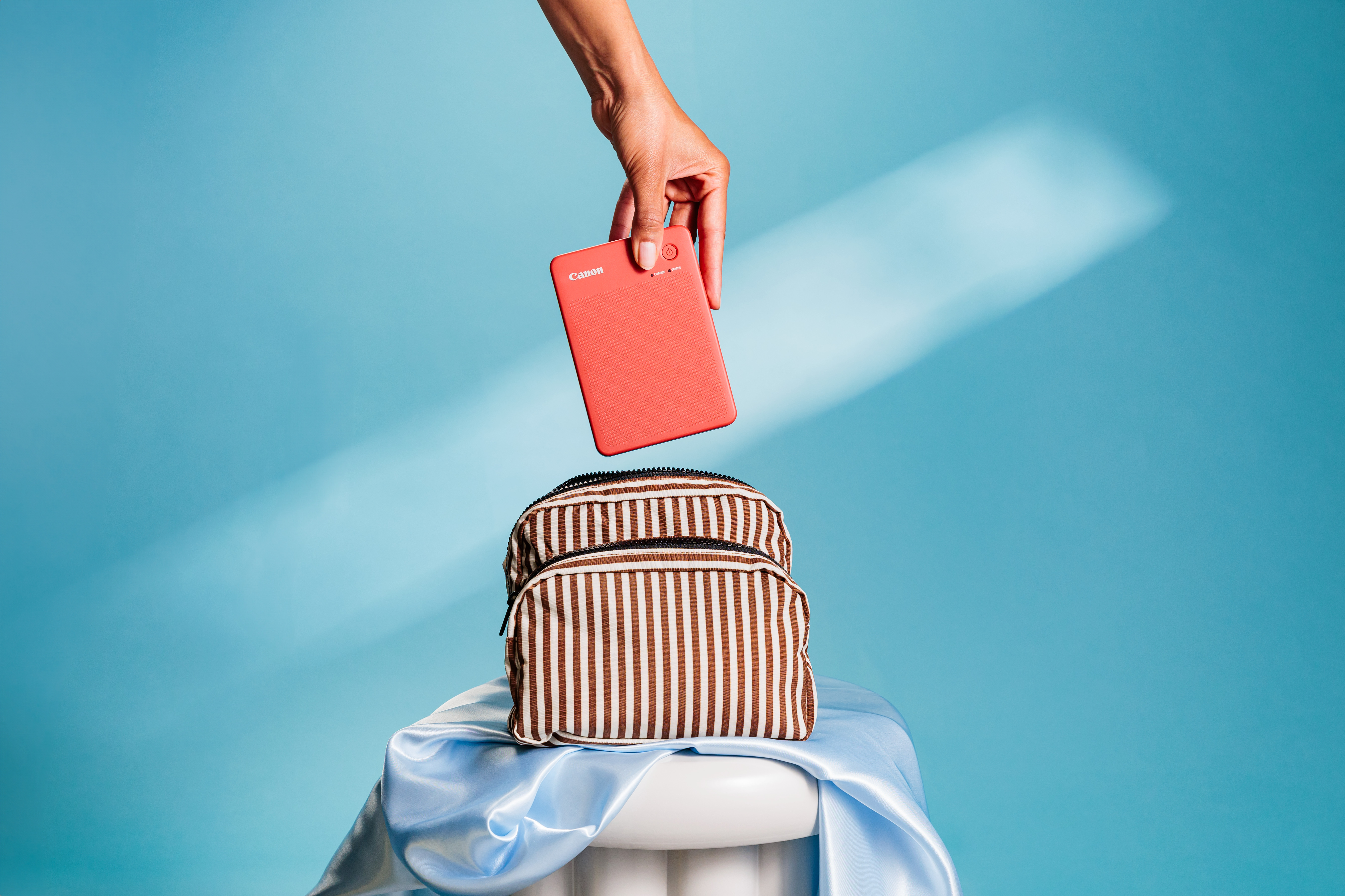 Eine Hand hält einen roten Drucker über einer gestreiften Tasche auf einem Sockel. Der Hintergrund ist ein blauer Farbverlauf.