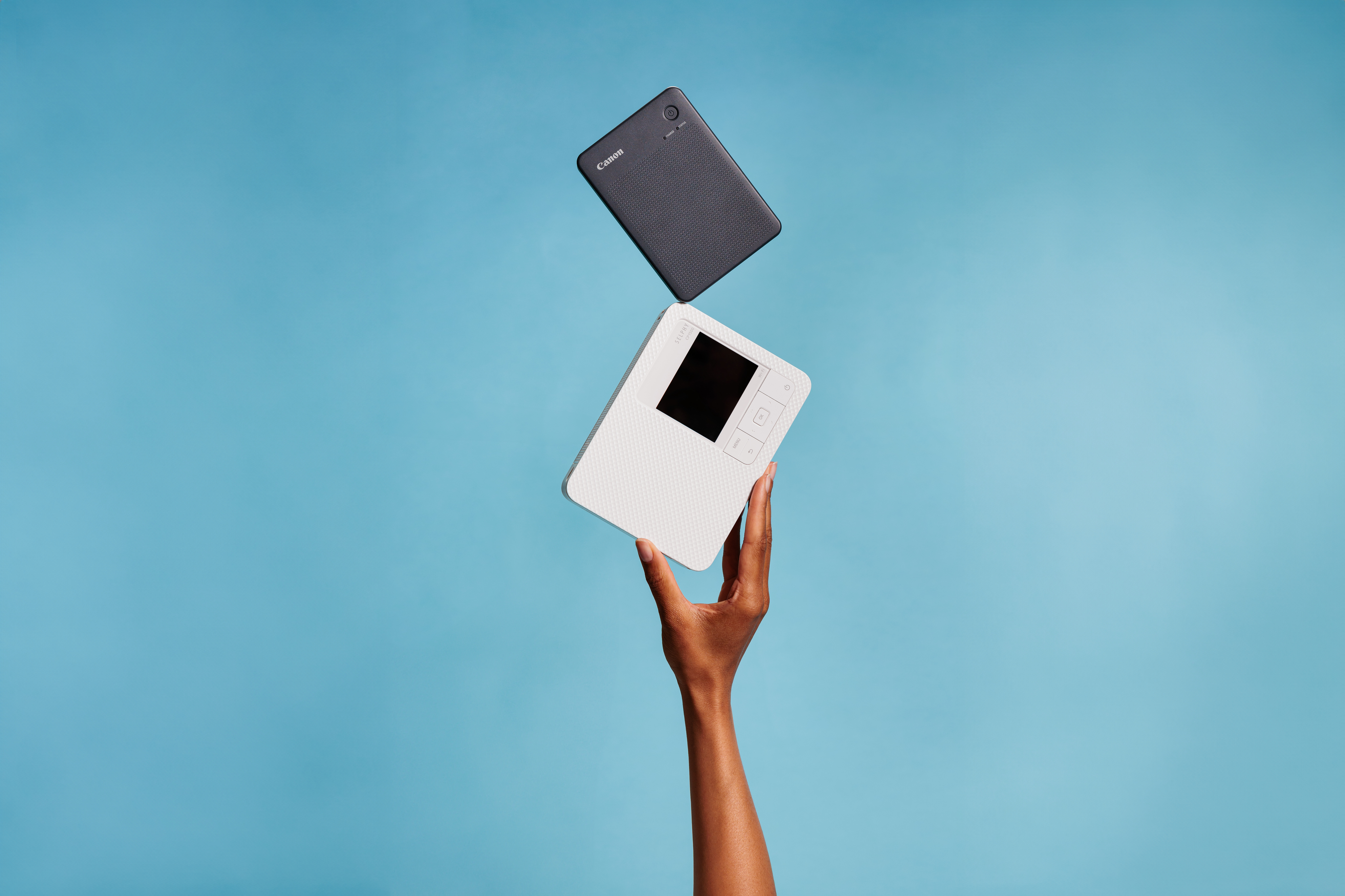 Eine Hand hält einen schwarzen und einen weißen Drucker über einem blauen Hintergrund. Die Drucker sind von Canon.
