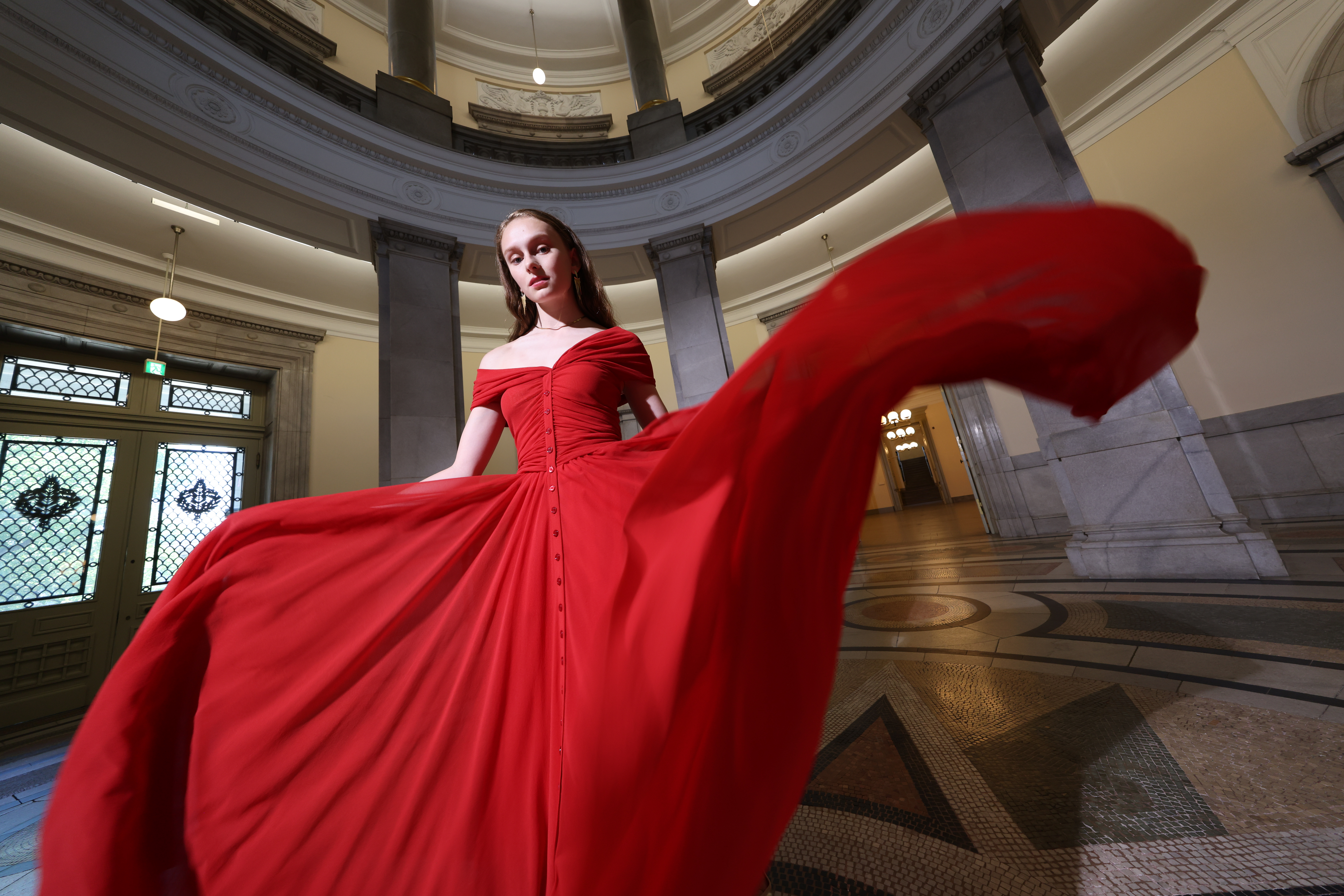 Frau in rotem Kleid, das sich in einem Gebäude mit Säulen und Gewölbedecke dreht.