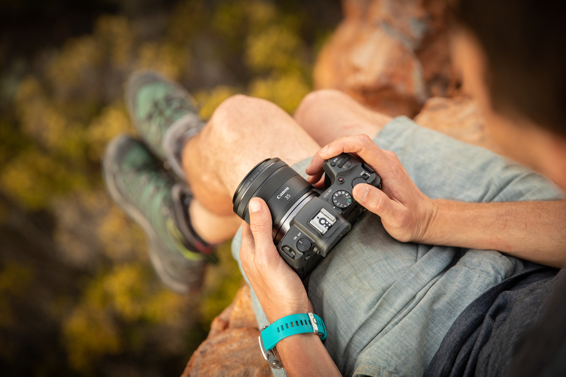 Eine Person hält eine schwarze Canon Kamera mit Objektiv und sitzt auf einem Felsen.