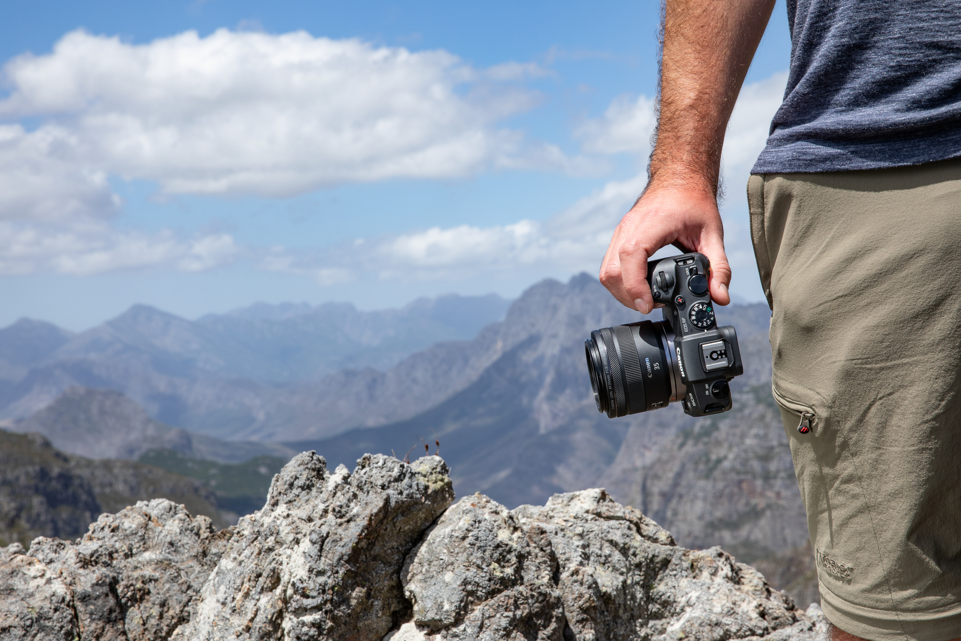 Eine Person hält eine schwarze Canon Kamera mit Objektiv, auf einem Berggipfel.