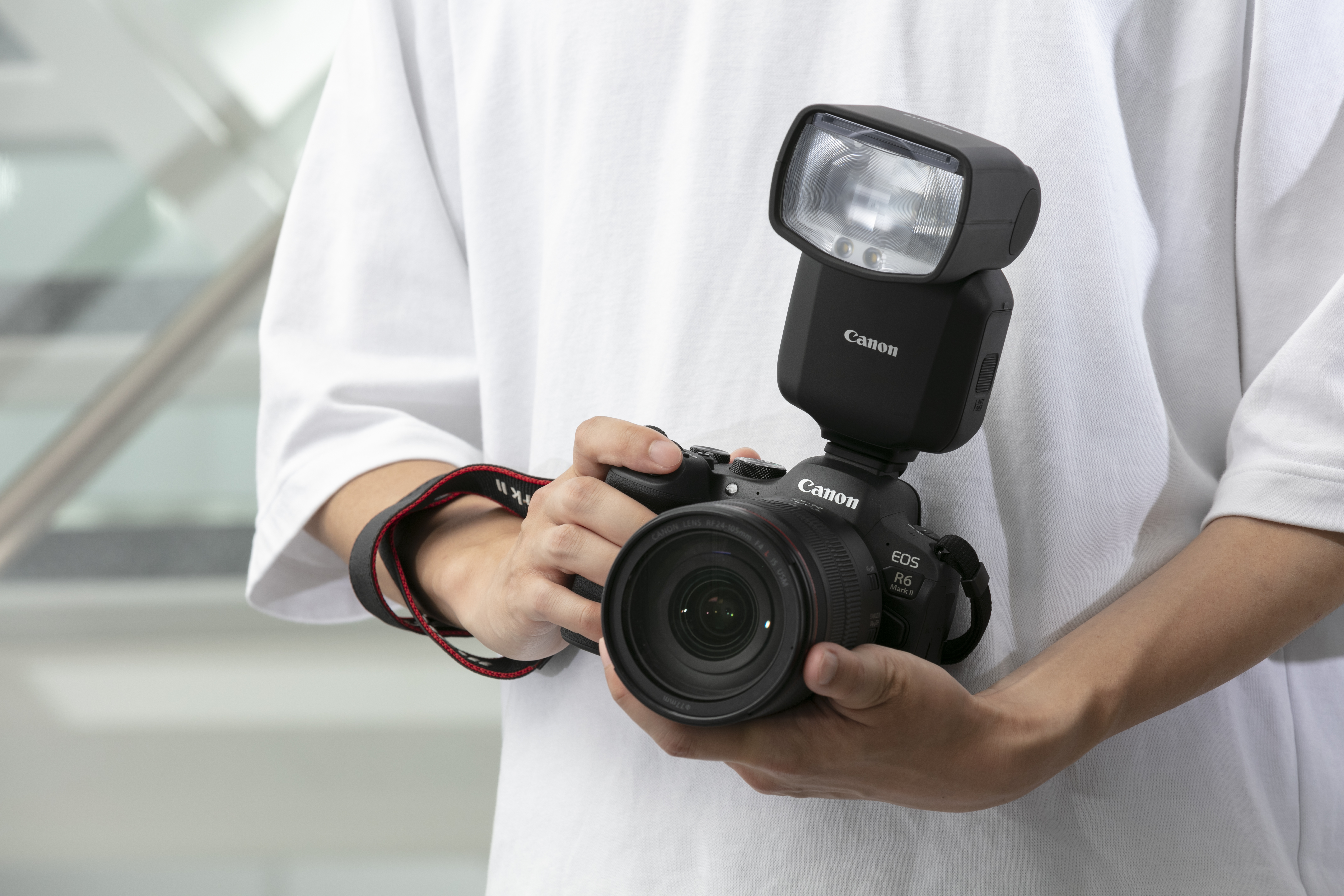 Person hält eine Canon-Kamera mit Blitz. Die Person trägt ein weißes Hemd. Der Hintergrund ist unscharf.